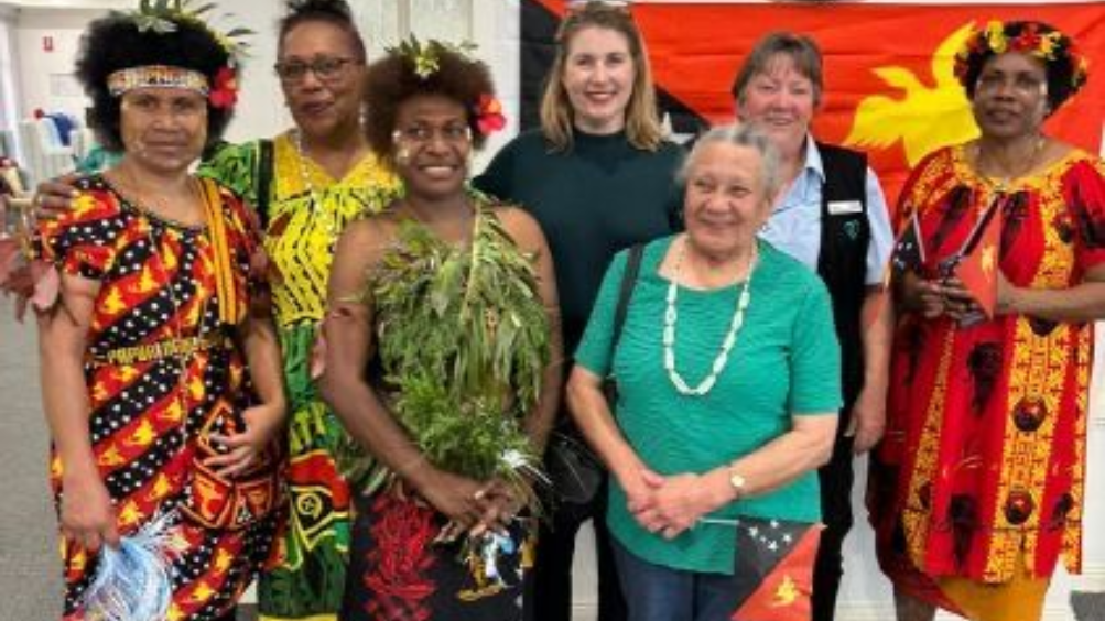 Group of women in Papua New Guinean attire pose with two women, indoors near a PNG flag.