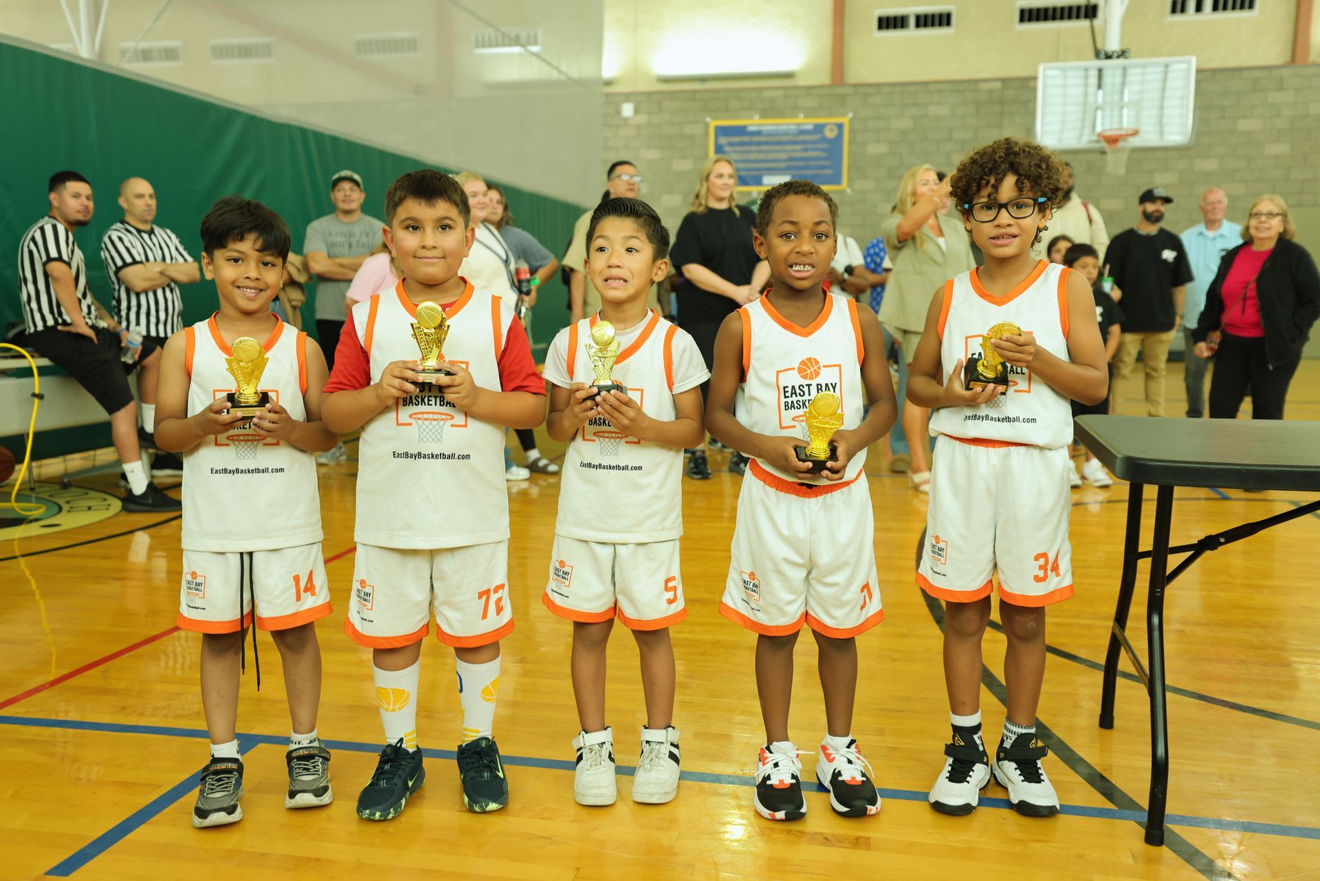 Five children in white and orange basketball uniforms hold small gold trophies while standing on a gymnasium floor.