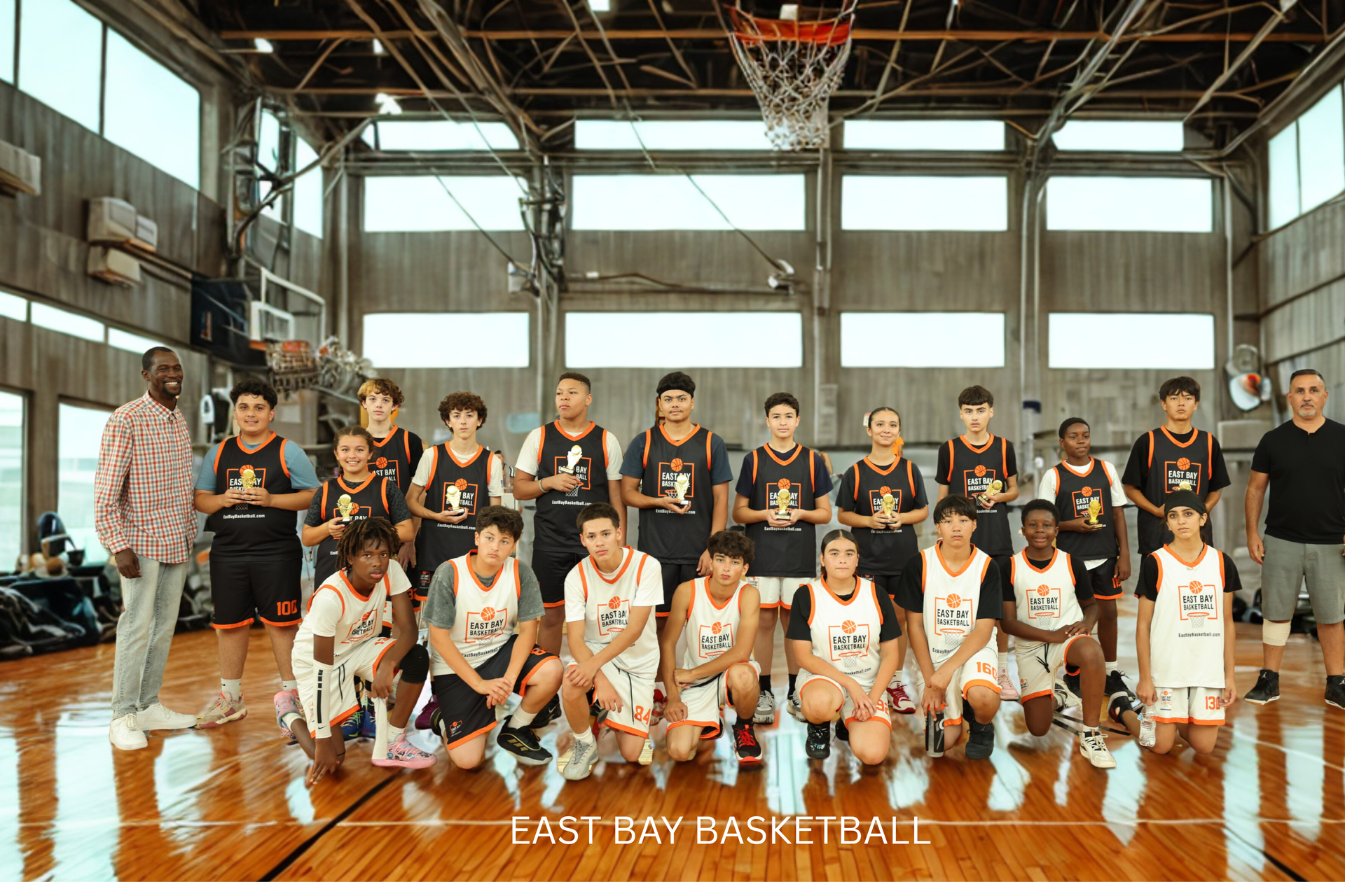 A basketball team poses for a group photo on a wooden indoor court at the East Bay Basketball training center.