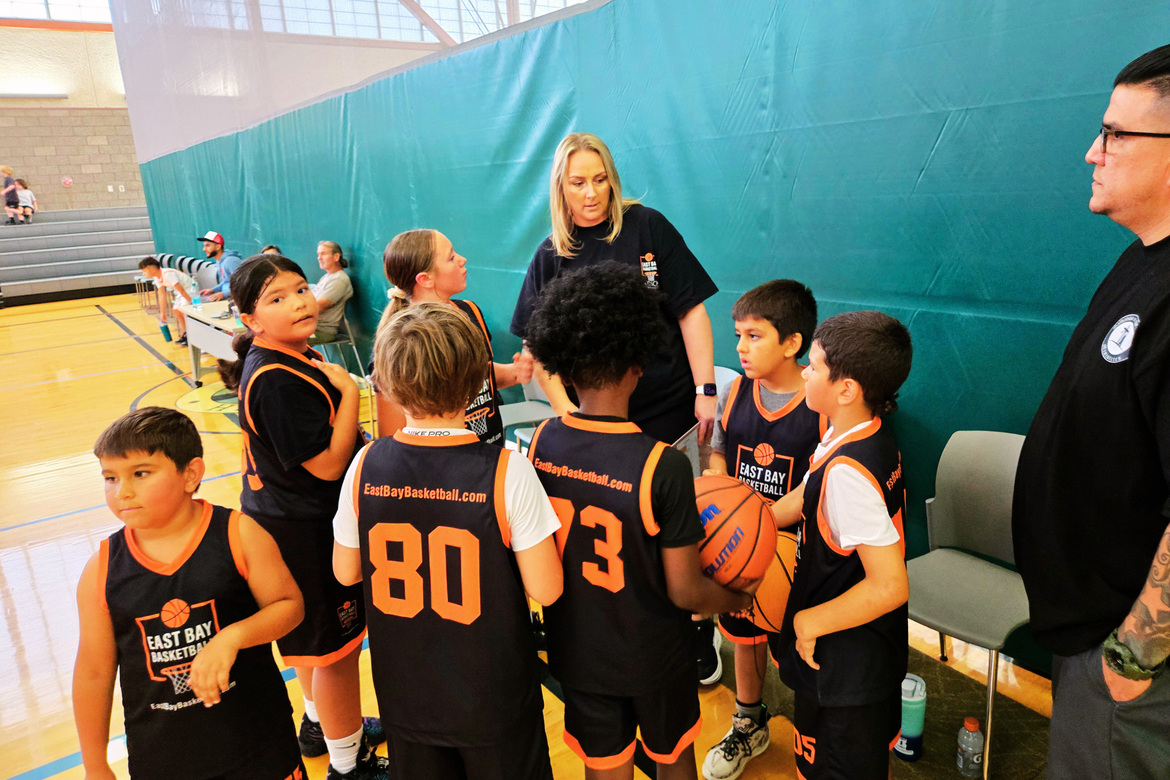 A coach gives instructions to a group of children in black and orange basketball jerseys on an indoor court.