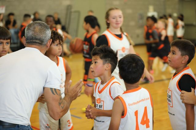 A coach speaks to a group of children in white and orange basketball jerseys on an indoor court.