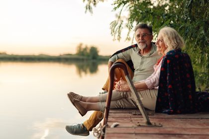 Two people sit on a wooden dock by a calm lake at sunset, relaxing and talking.