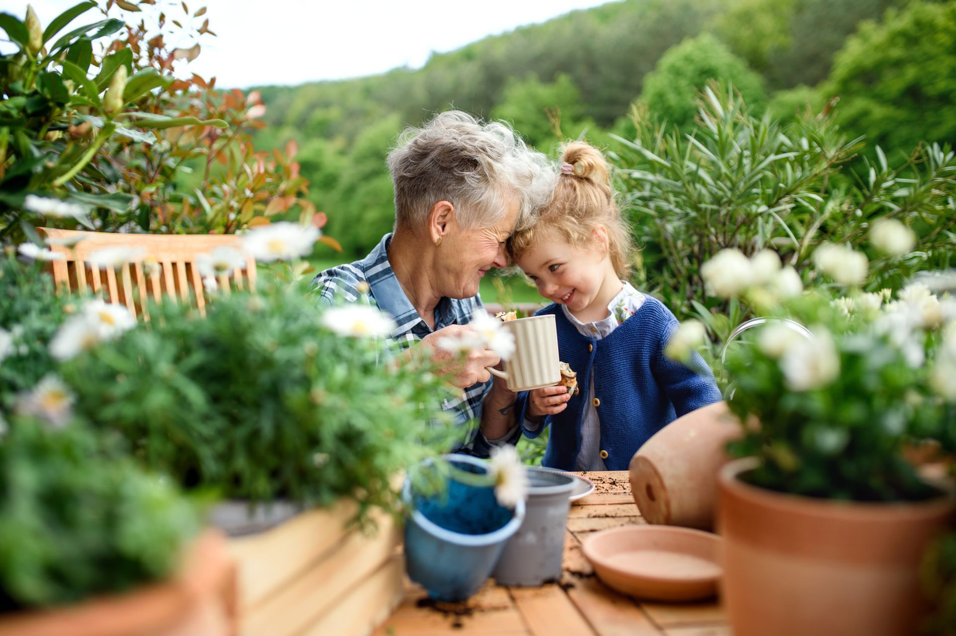 Two people smiling over mugs in a lush garden with potted plants and flowers