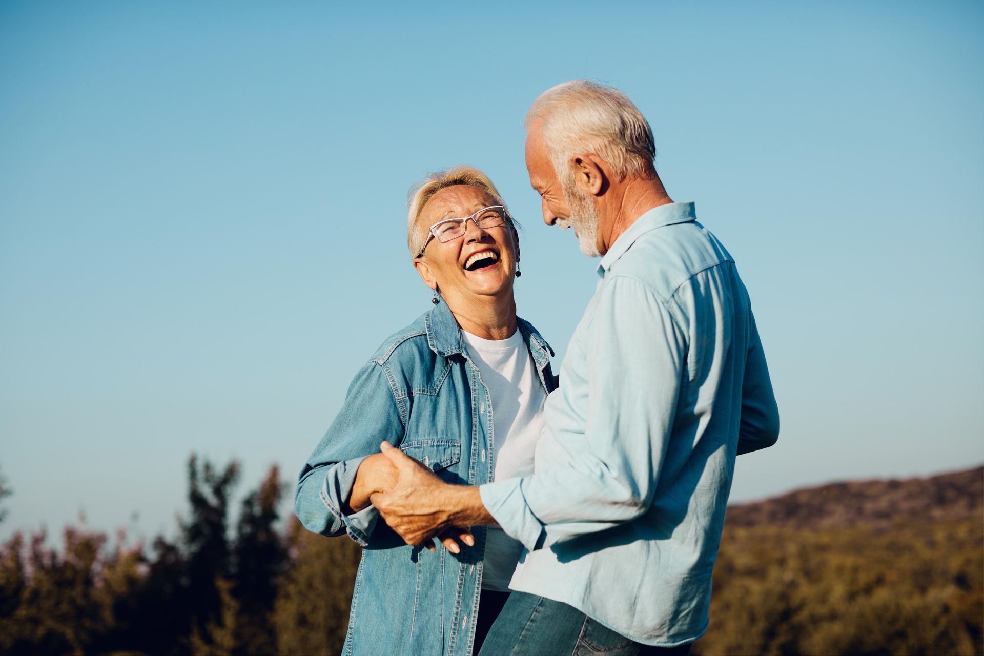 Two people laughing and holding hands in a sunlit field under a clear blue sky