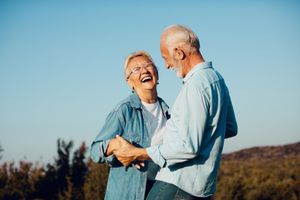 Two people laughing and holding hands in a sunlit field under a clear blue sky