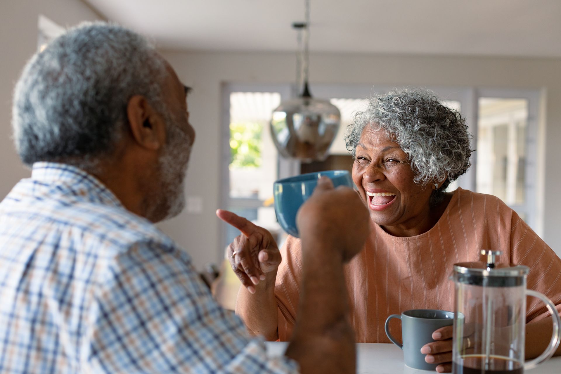 Two people smiling and talking at a kitchen table with mugs and a French press
