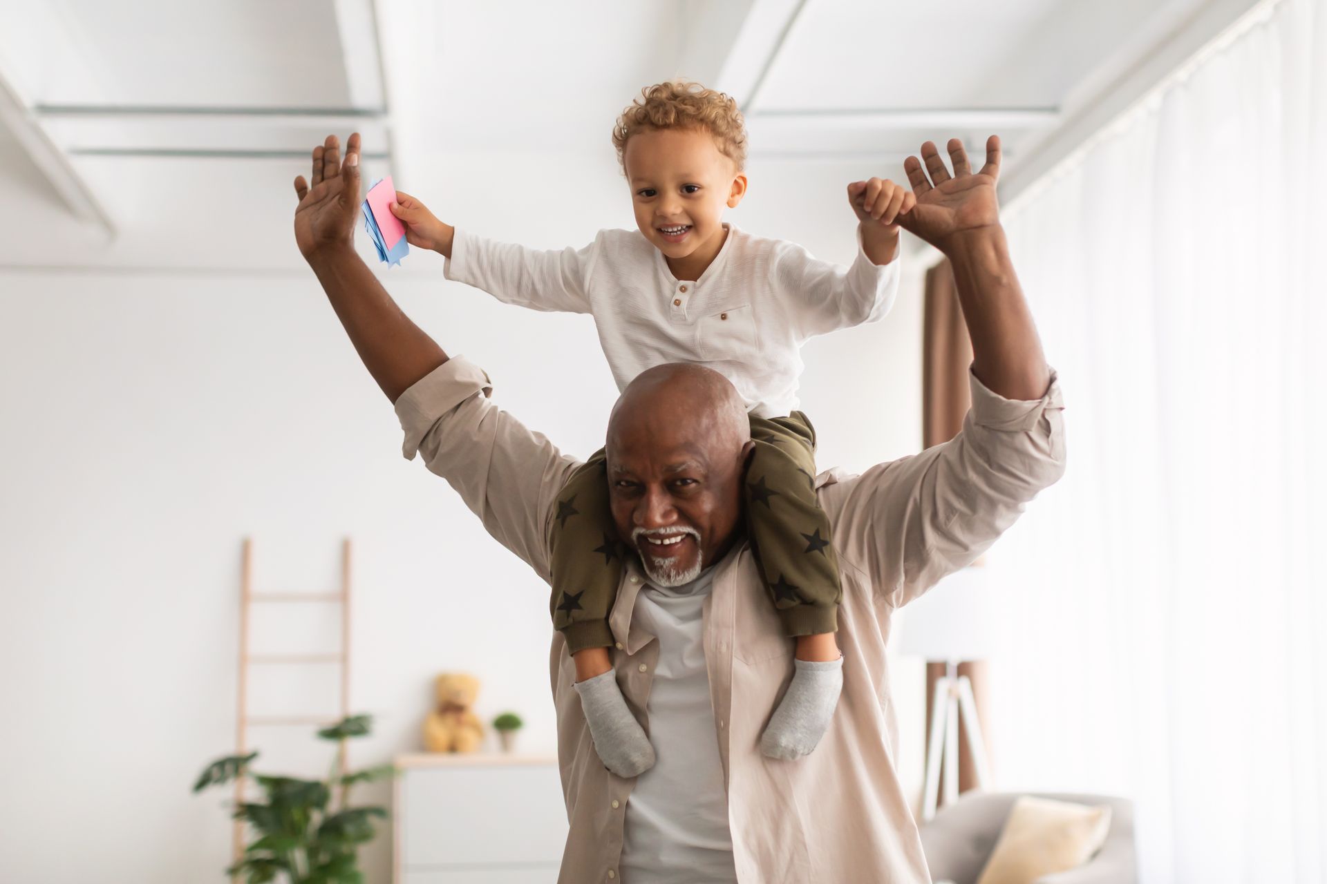 Adult carrying smiling child on shoulders in a bright living room, both with arms raised