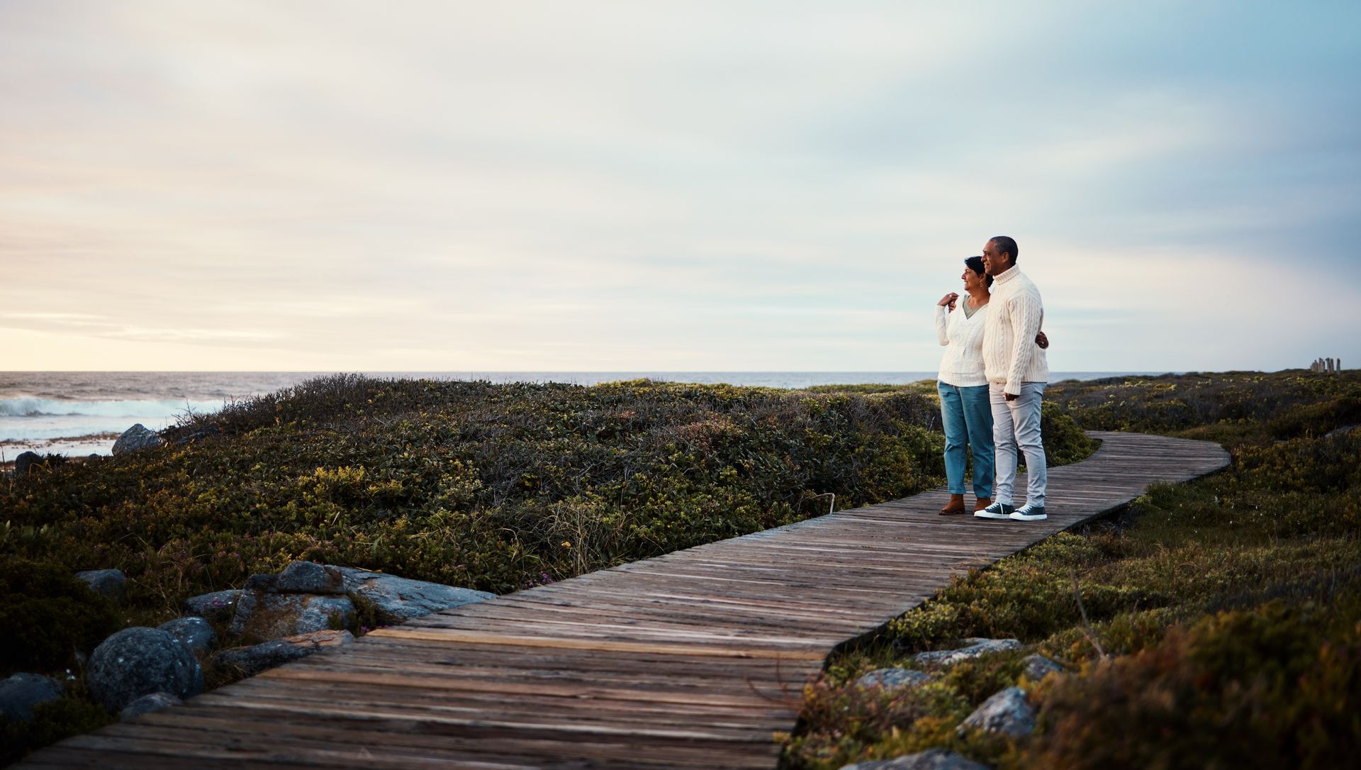 Two people standing on a wooden boardwalk by the sea at sunset