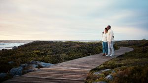 Two people standing on a wooden boardwalk by the sea at sunset