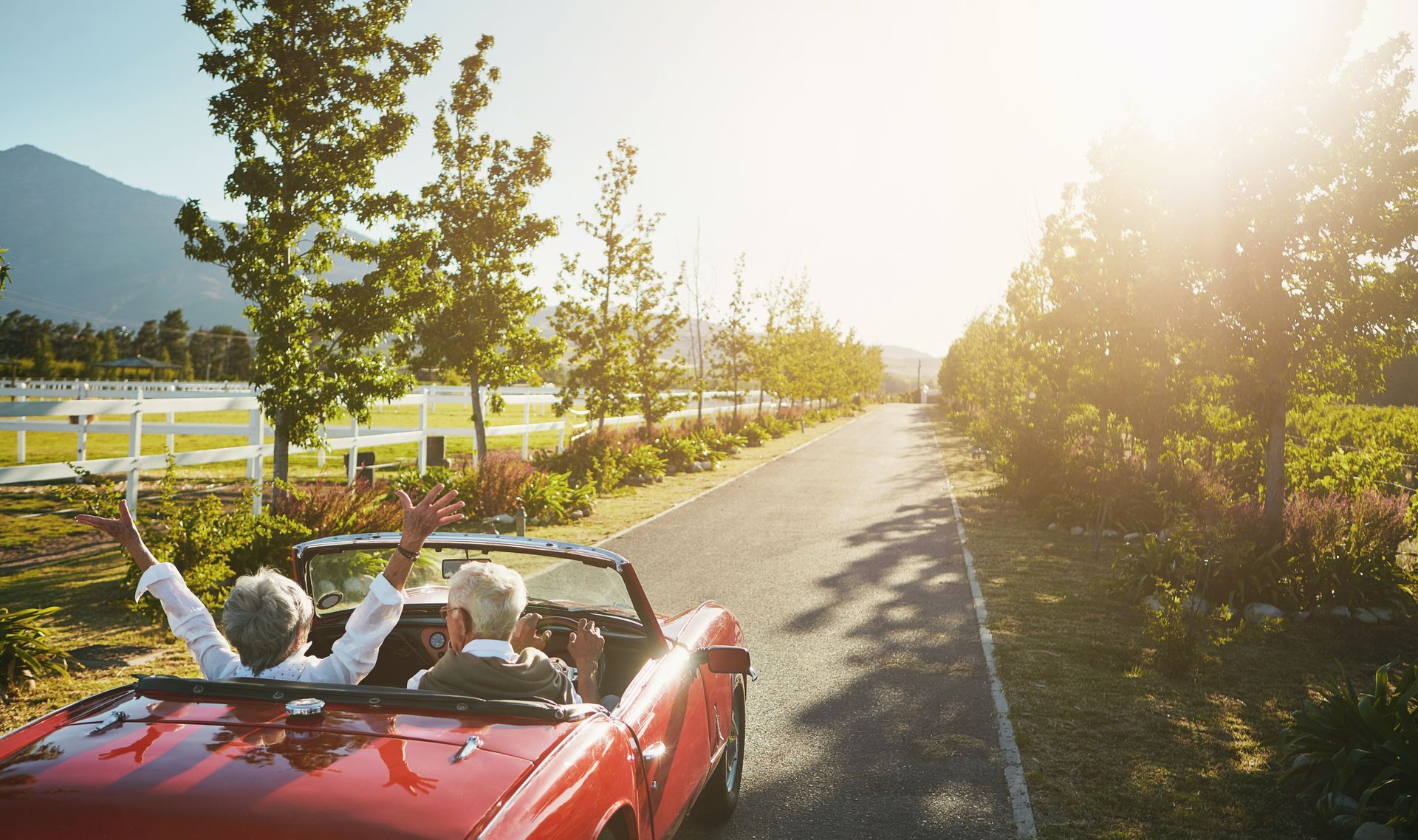 Red convertible driving on a sunny country road, with white flowers and trees along the fence.