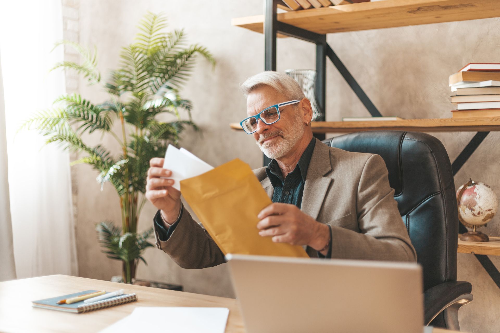 Man in blazer opening envelope at a desk, looking at papers, smiling. Bookshelf, laptop, and plant in the background.