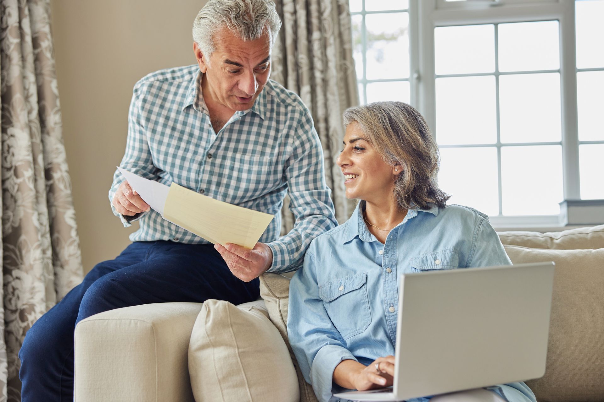 Couple reviewing paperwork together while using a laptop on a couch in a home.