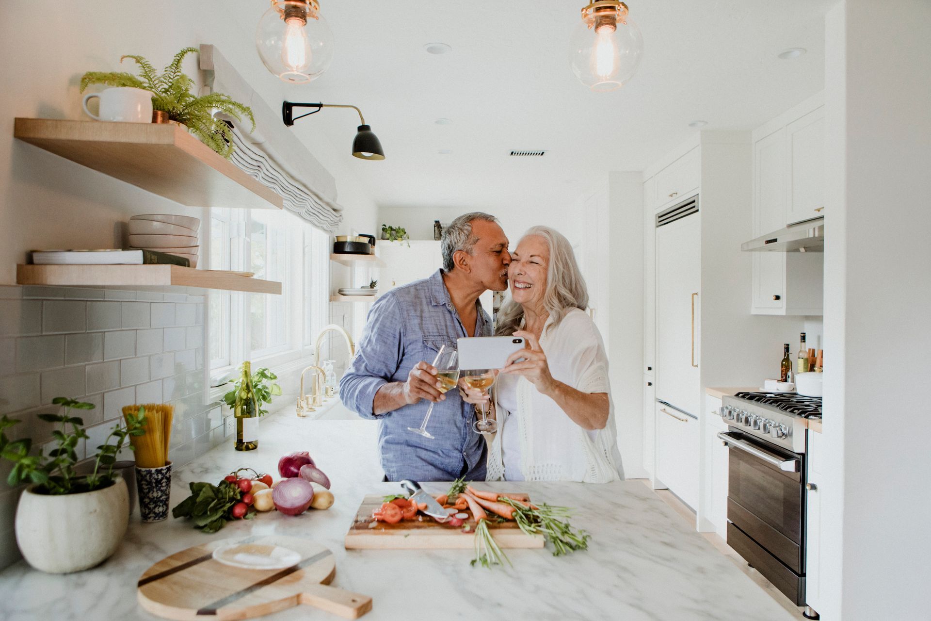 Senior couple in kitchen: man kissing woman's cheek, holding wine glasses, preparing food.