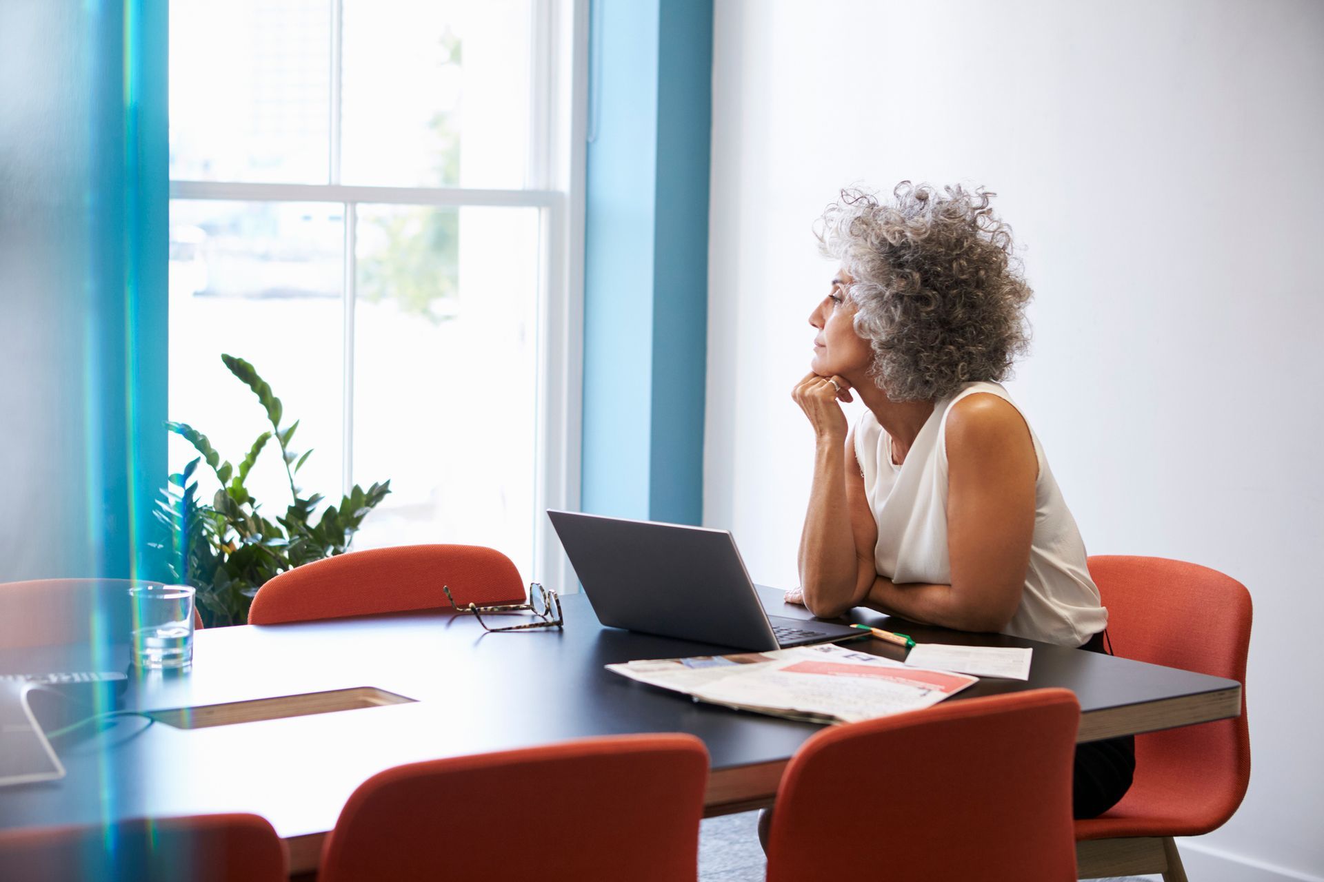 Woman with curly gray hair, at a conference table, gazing out a window, hand to her chin, laptop open.