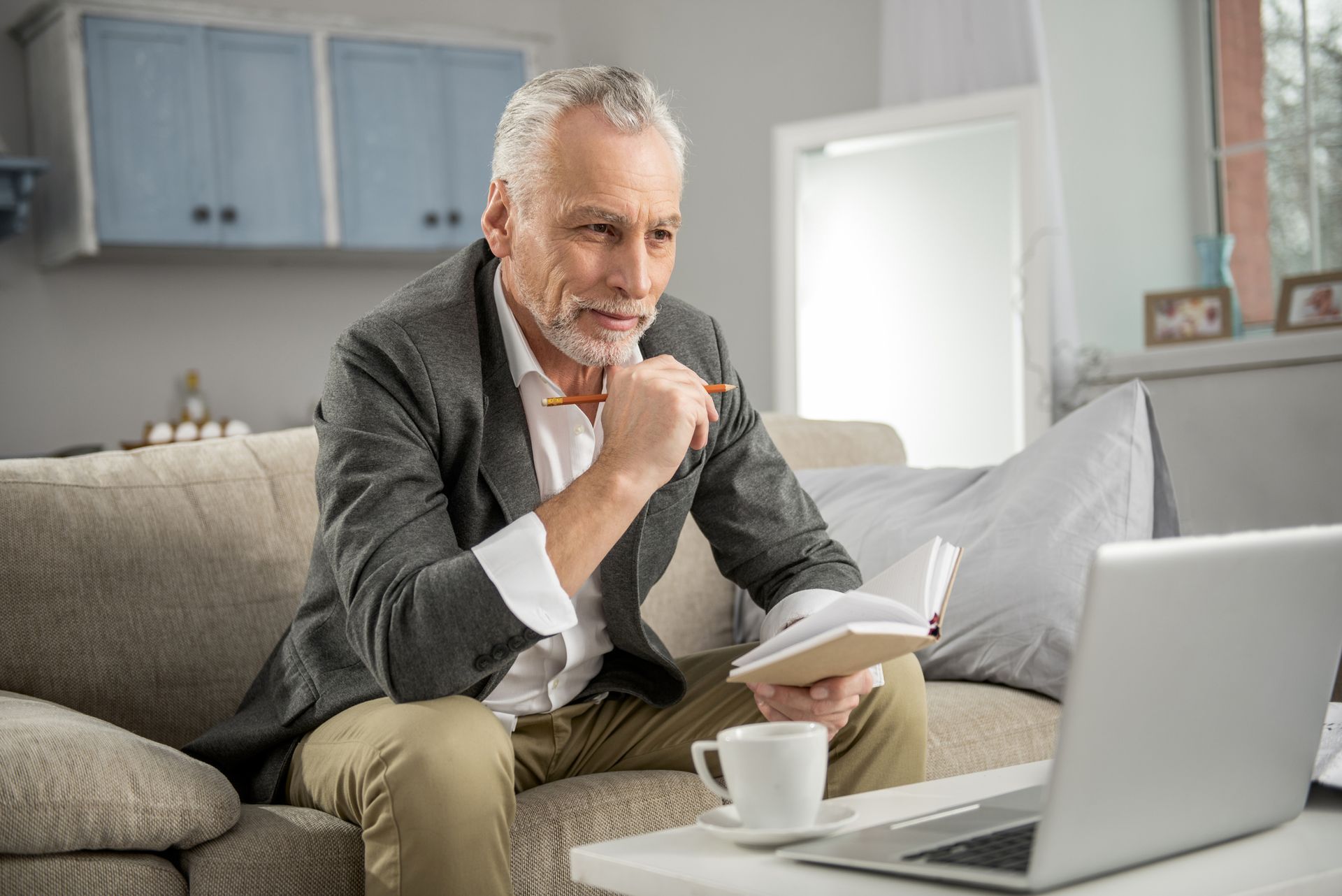 Man with gray hair sits on couch, looking at laptop, holding notebook and pen. Coffee on table.