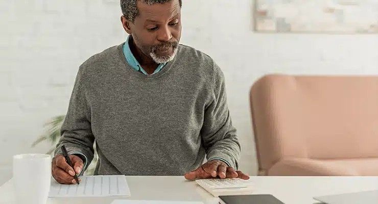 An older Black man with a gray beard, writing on paper and using a calculator at a table.