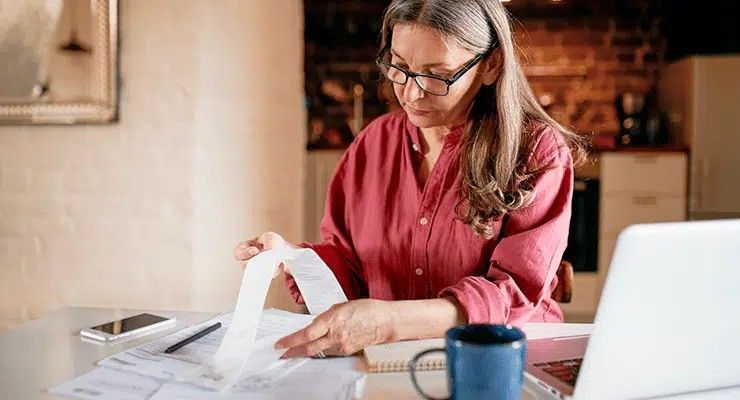 Woman with glasses reviews bills at a table with a laptop, phone, and coffee mug.