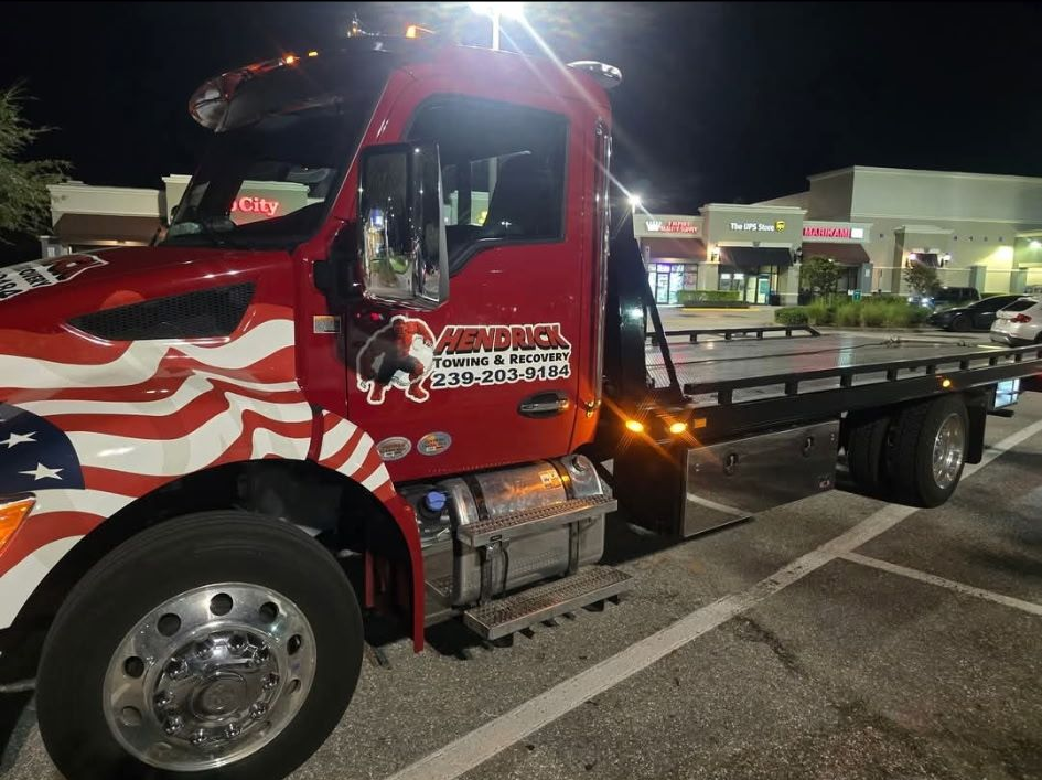Red tow truck with American flag design parked at night in a lot with storefronts.