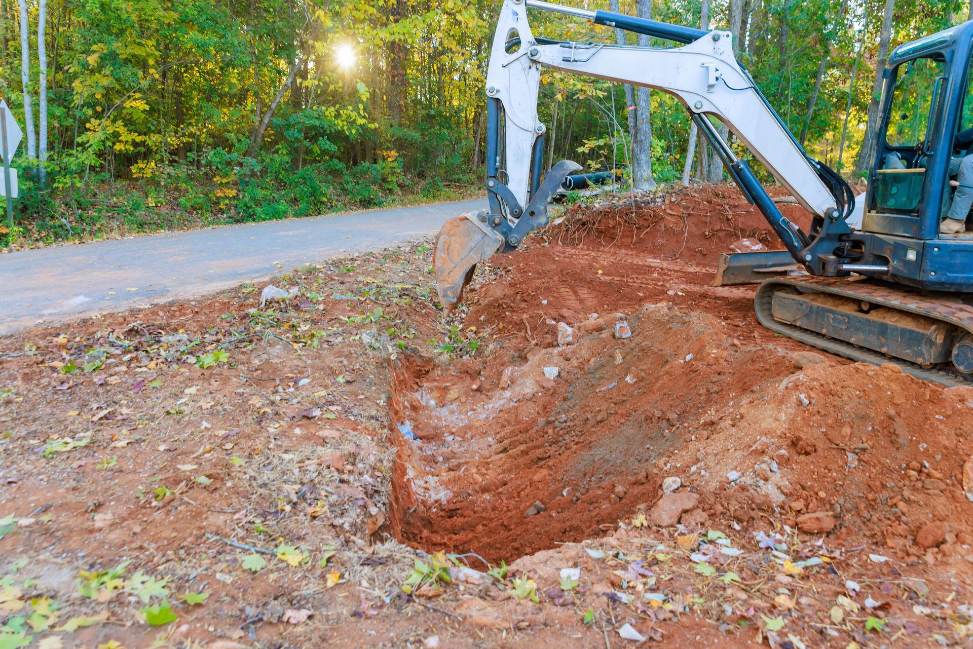 Mini excavator digging a trench beside a road in a wooded area. Sunlight visible.