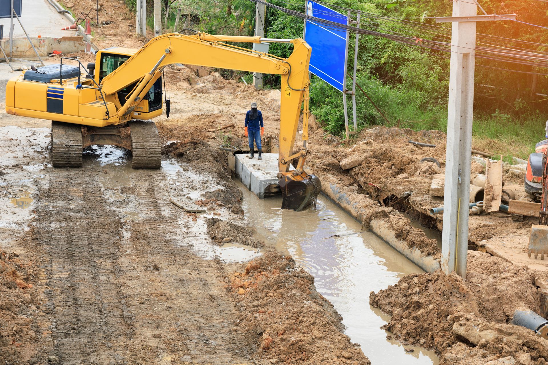 Yellow excavator digging in muddy trench; worker stands nearby.