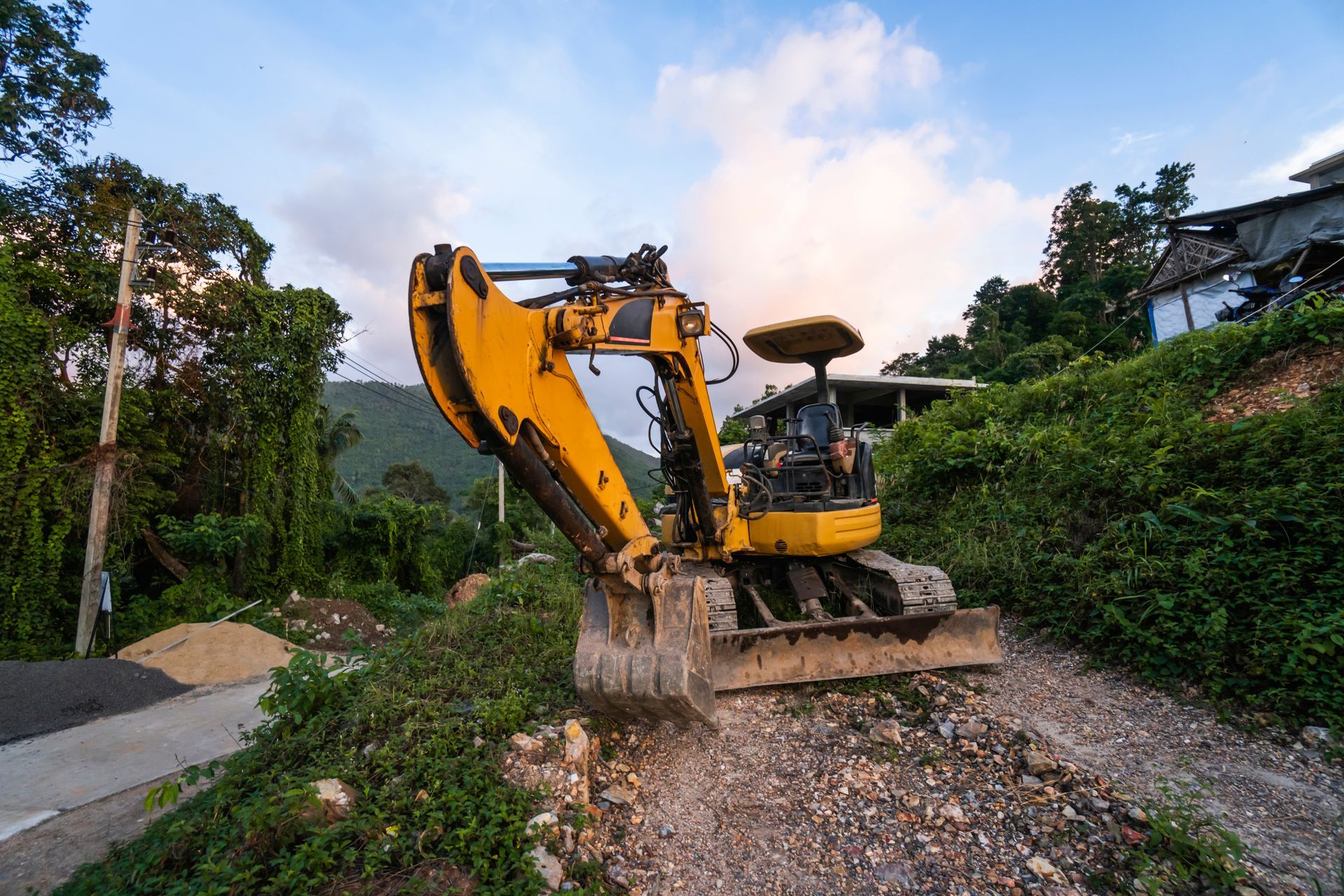 Yellow excavator on a rocky hill clearing land for construction with green foliage.