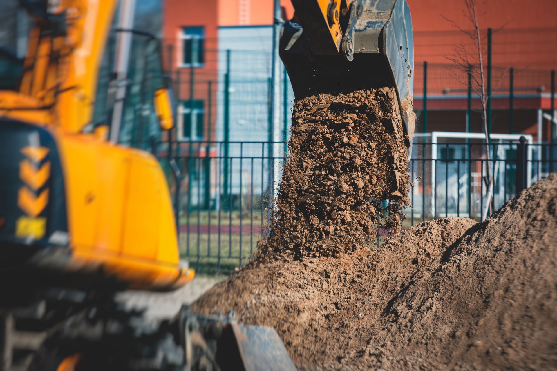 Yellow excavator dumping dirt near a building and fence.