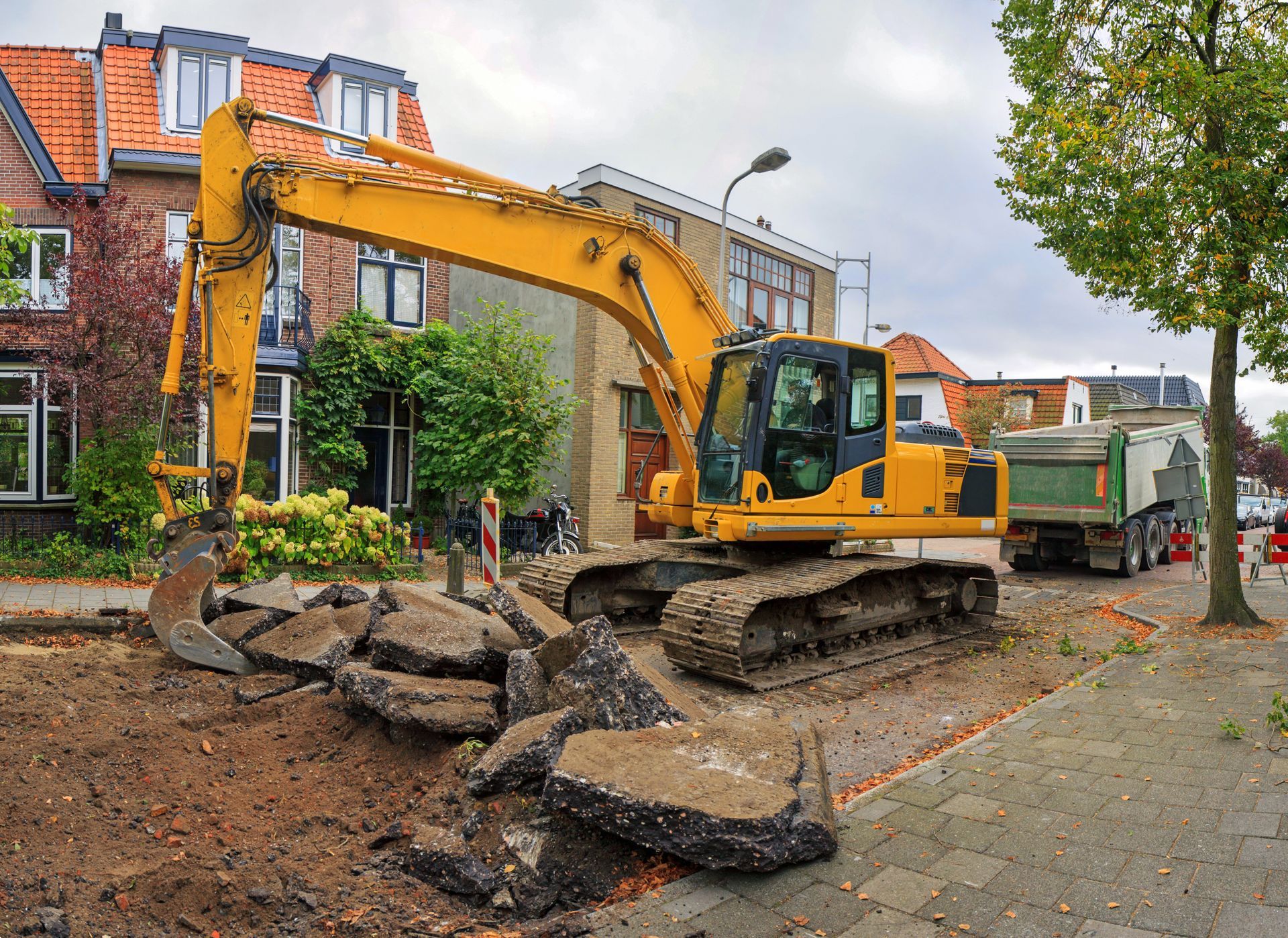Yellow excavator tearing up asphalt road in residential area.