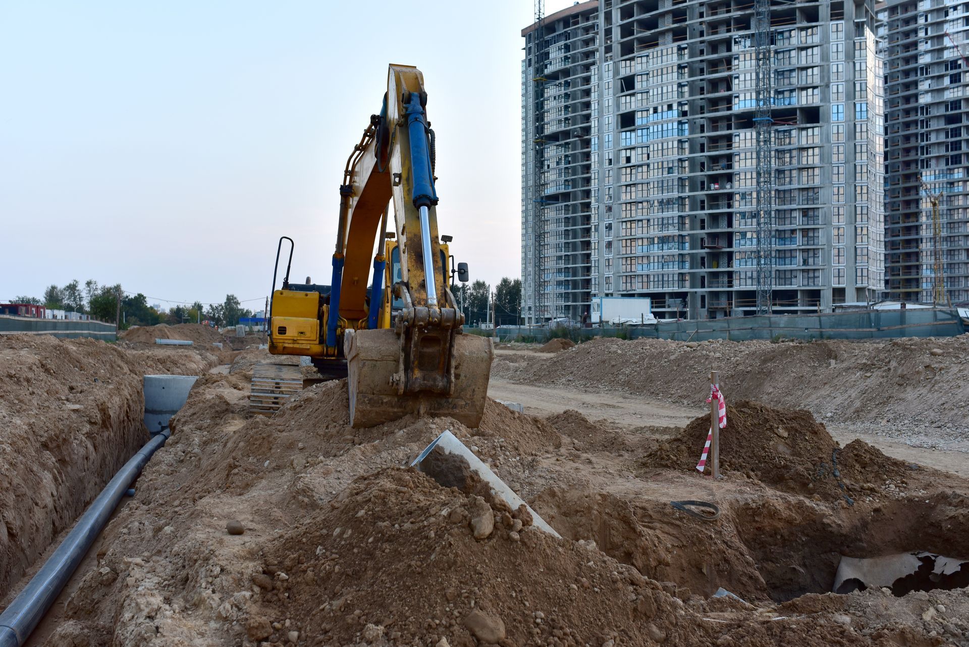 Excavator digging trench at construction site with tall buildings in the background.