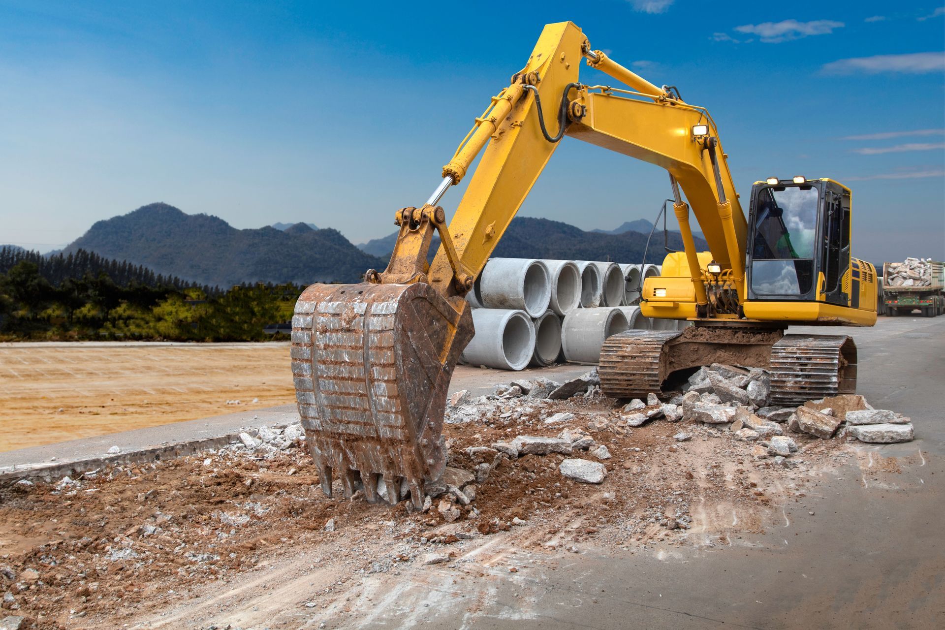 Yellow excavator demolishing pavement at a construction site with mountains in the background.
