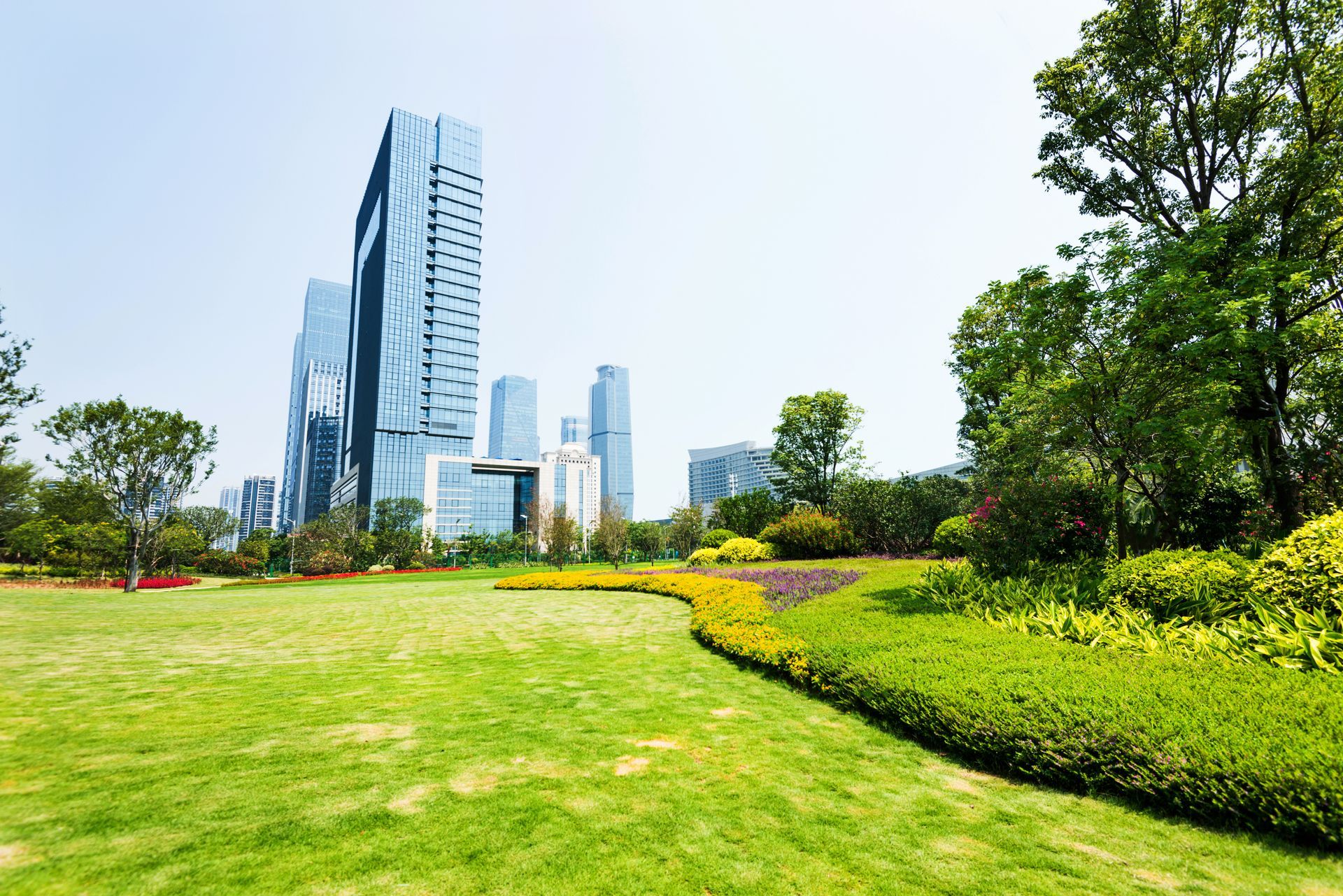 Green park with manicured lawns, flower beds, and modern skyscrapers in the background under a blue sky. Green park with manicured lawns, flower beds, and modern skyscrapers in the background under a blue sky.