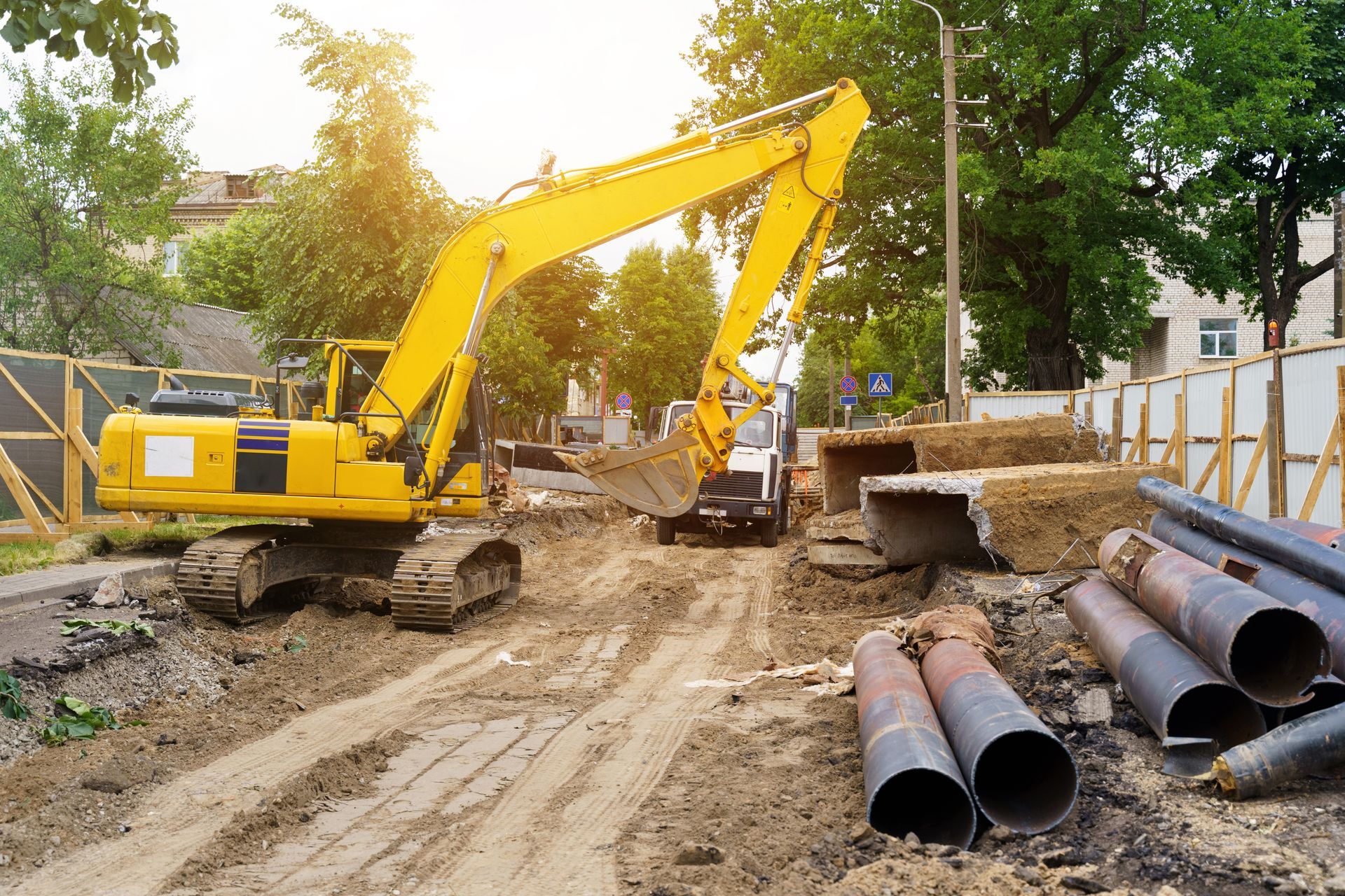 Yellow excavator digging on a construction site with pipes and a truck visible; sunny day.