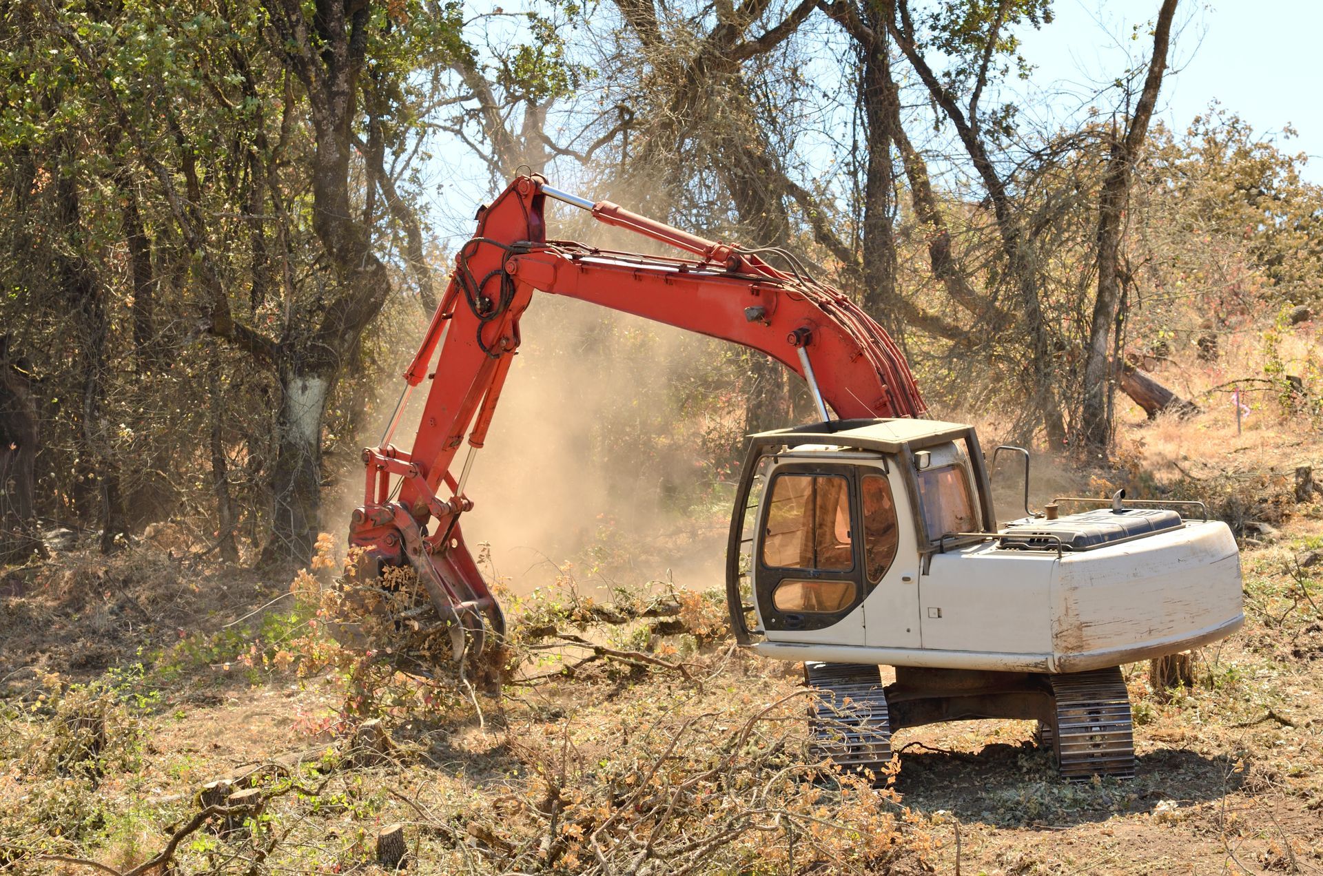 Red and white excavator clearing land, kicking up dust; trees in background.