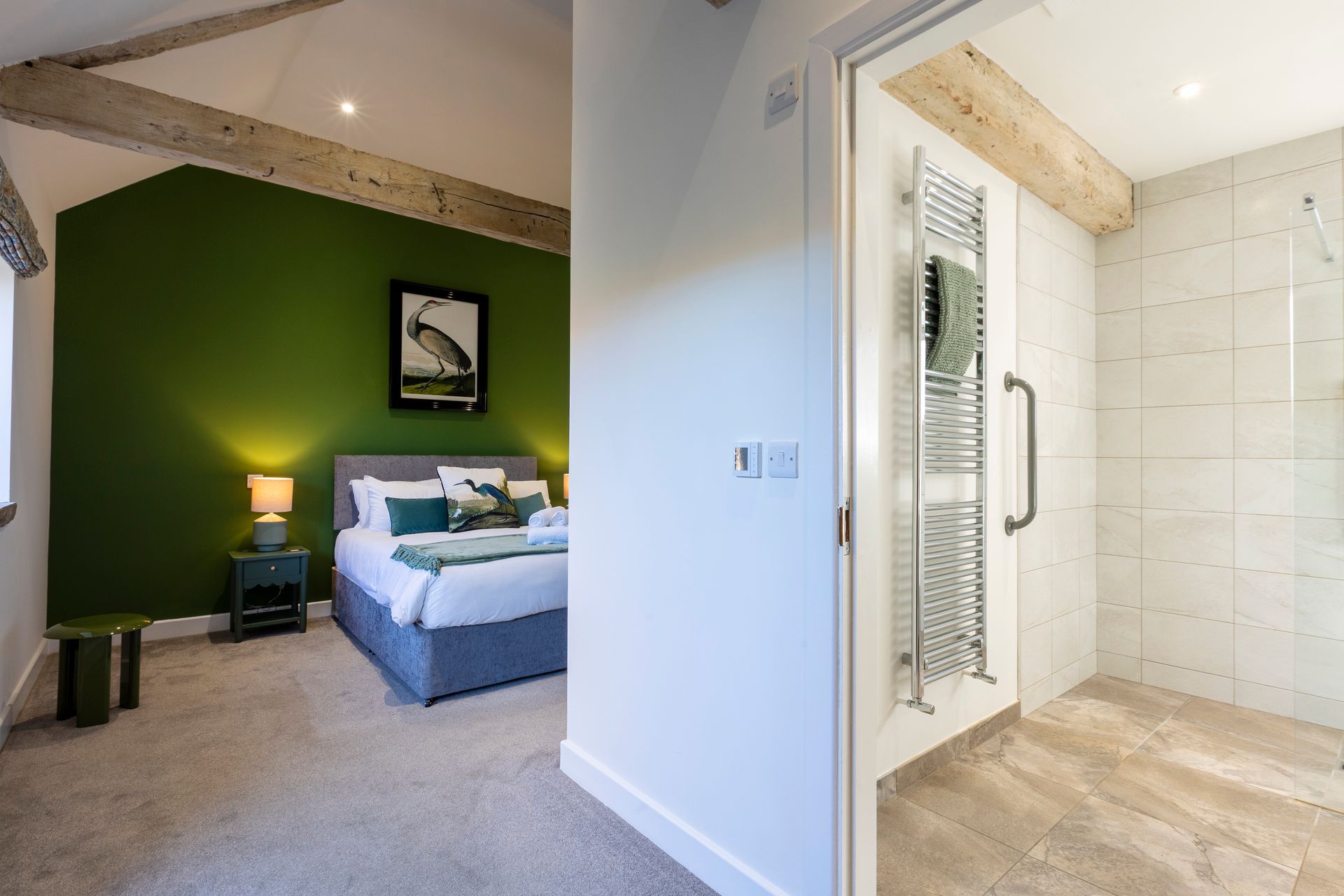 Bedroom with green accent wall, bed, and ensuite bathroom. Beige walls, wood beams.