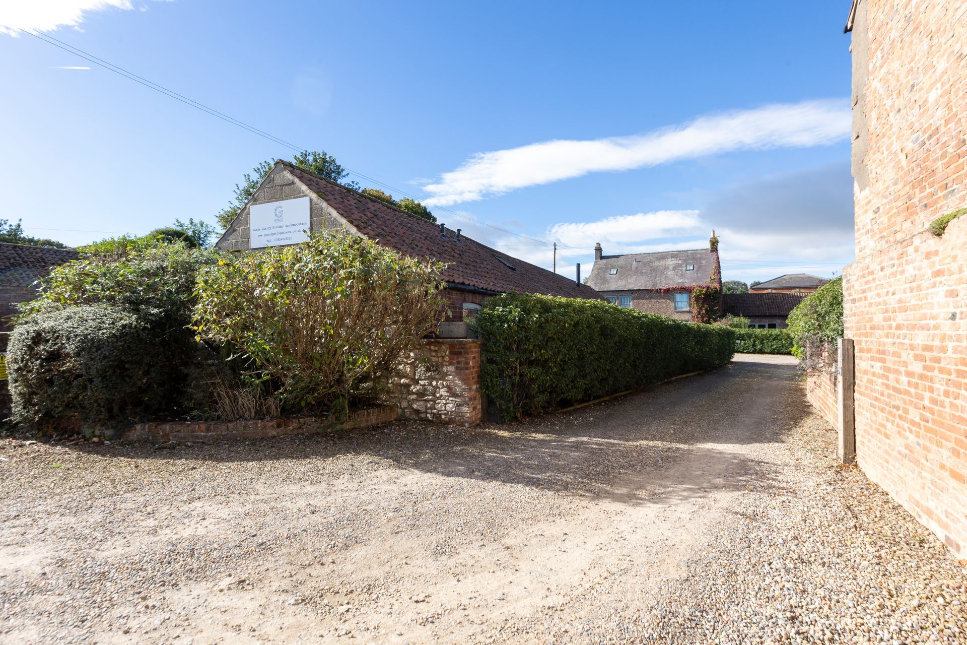 Gravel driveway leads toward buildings, a brick wall on the right and green hedge on the left, under blue sky.