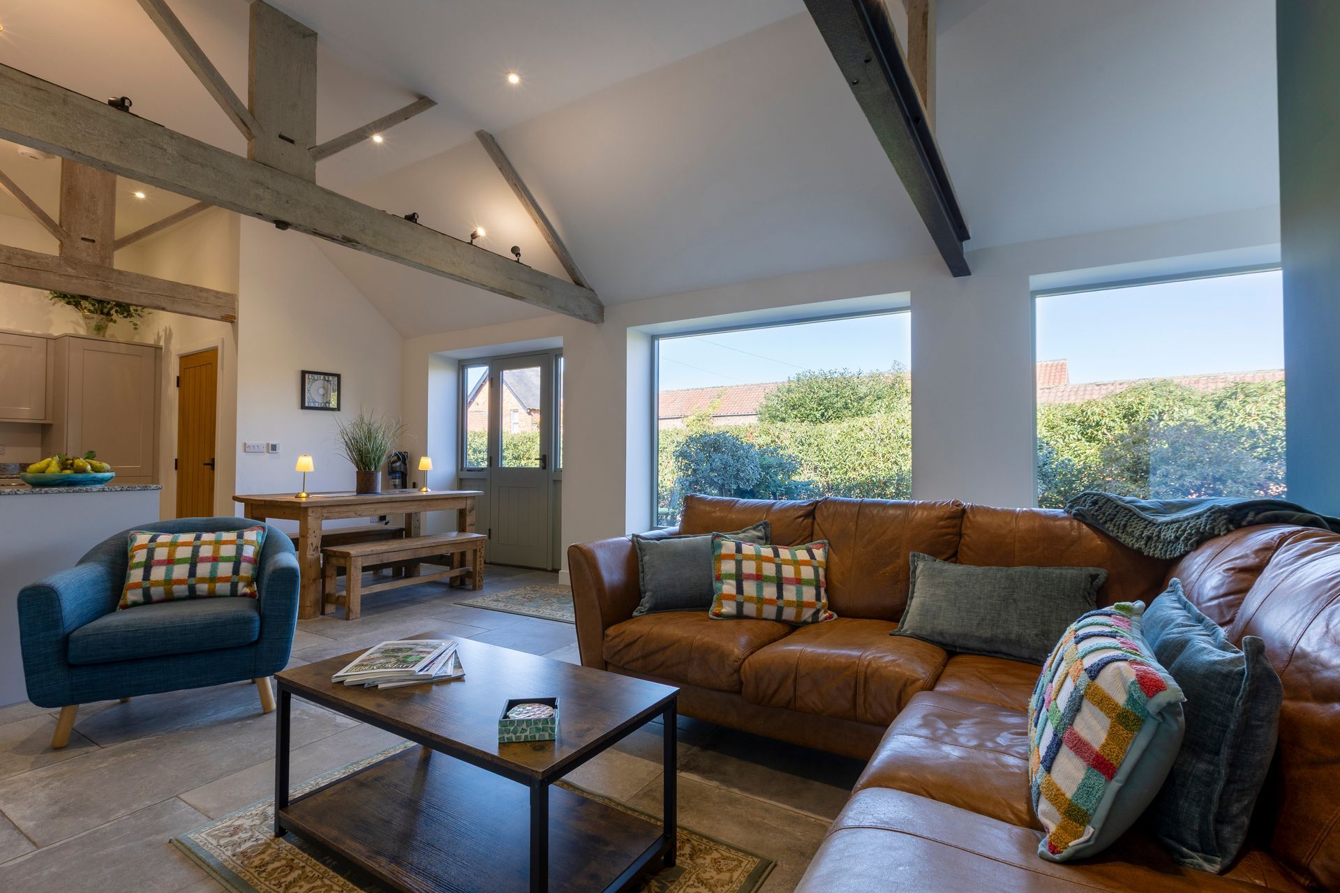 Living room with exposed beams, leather sofa, blue armchair, large windows overlooking greenery.