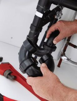 Des mains assemblent des tuyaux de plomberie noirs sous un évier. Une clé à molette se trouve à proximité.