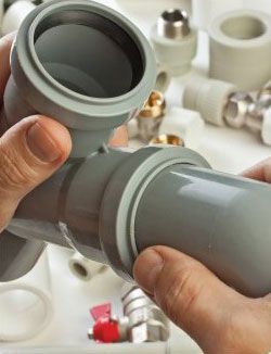 Des mains assemblent des tuyaux et des raccords de plomberie en plastique gris.