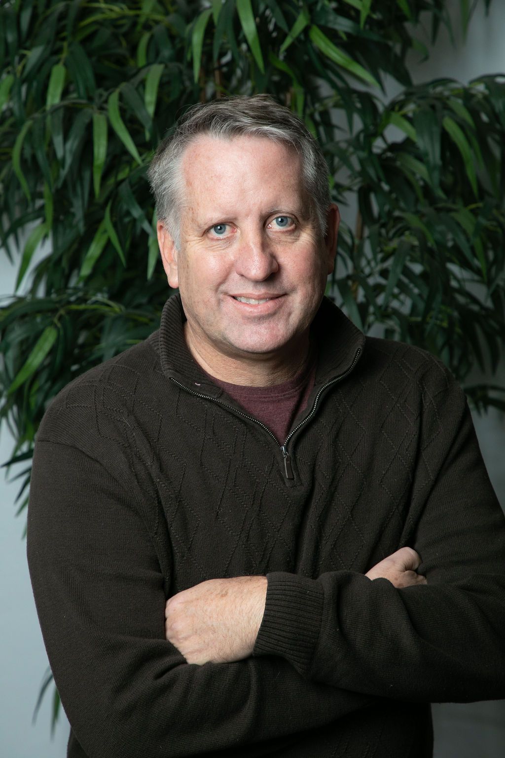 A man in a brown sweater is standing with his arms crossed in front of a plant.