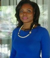A woman wearing a blue shirt and a pearl necklace is standing in front of a window.