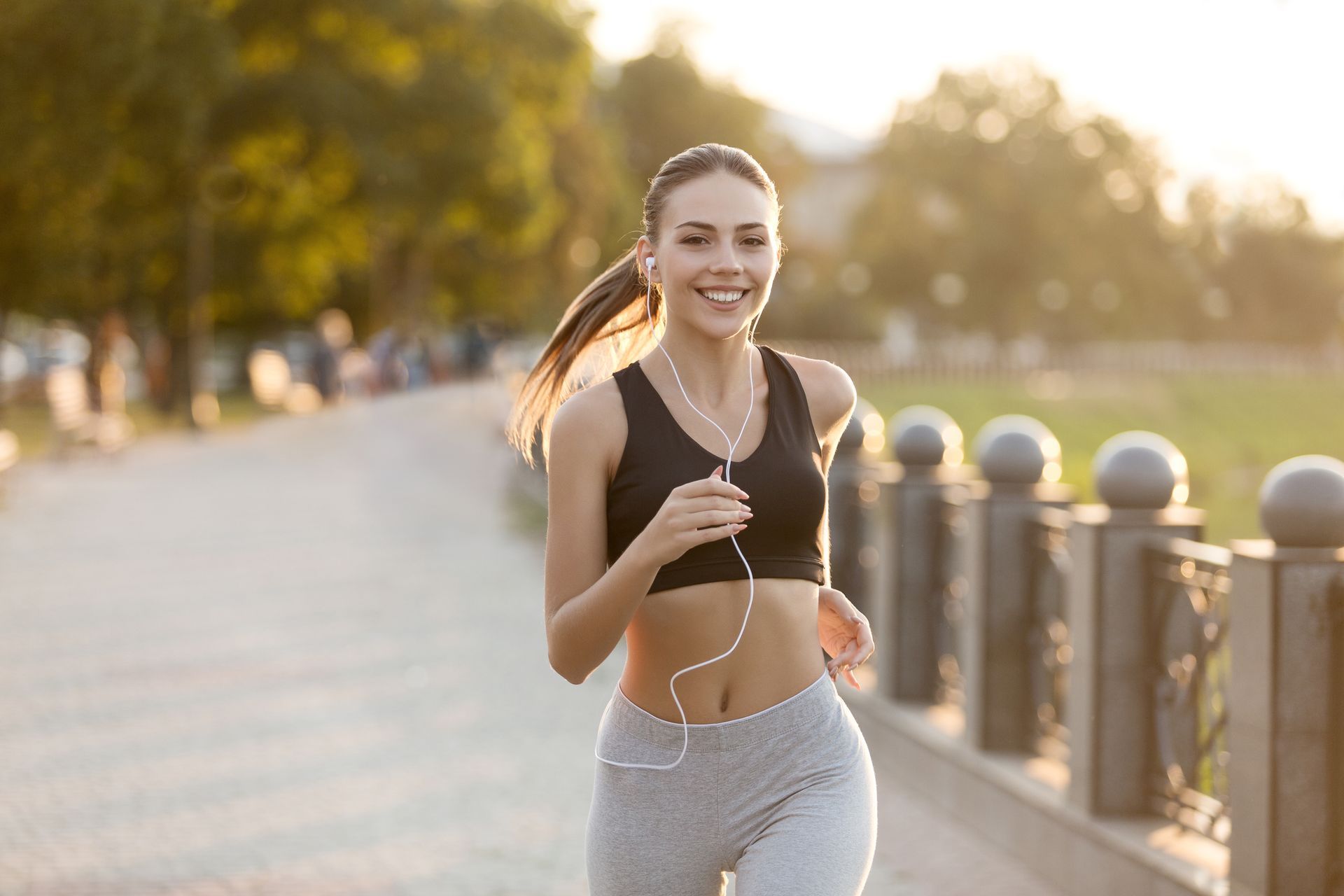 Woman running on a path, smiling, wearing a black sports bra, gray leggings, and earphones. Outdoors, sunny.