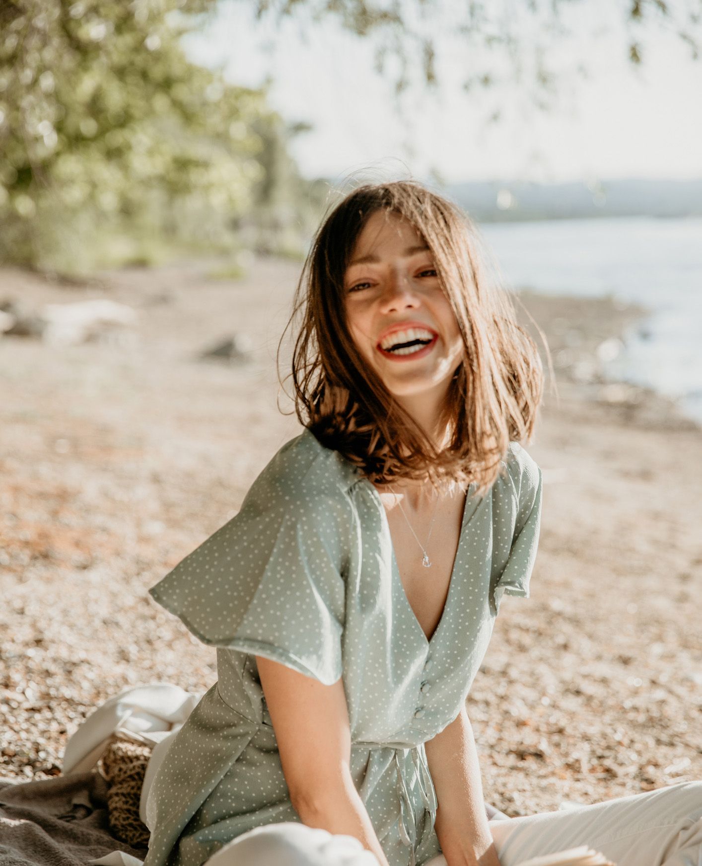 Woman laughing on a blanket near a lake, wearing a green polka dot dress.