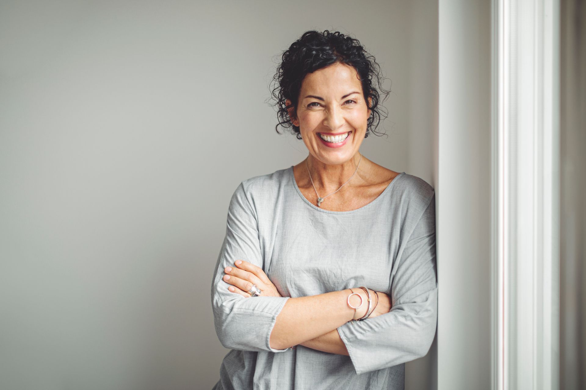 Woman with curly hair smiles, arms crossed, leaning against a wall. She wears a gray shirt.