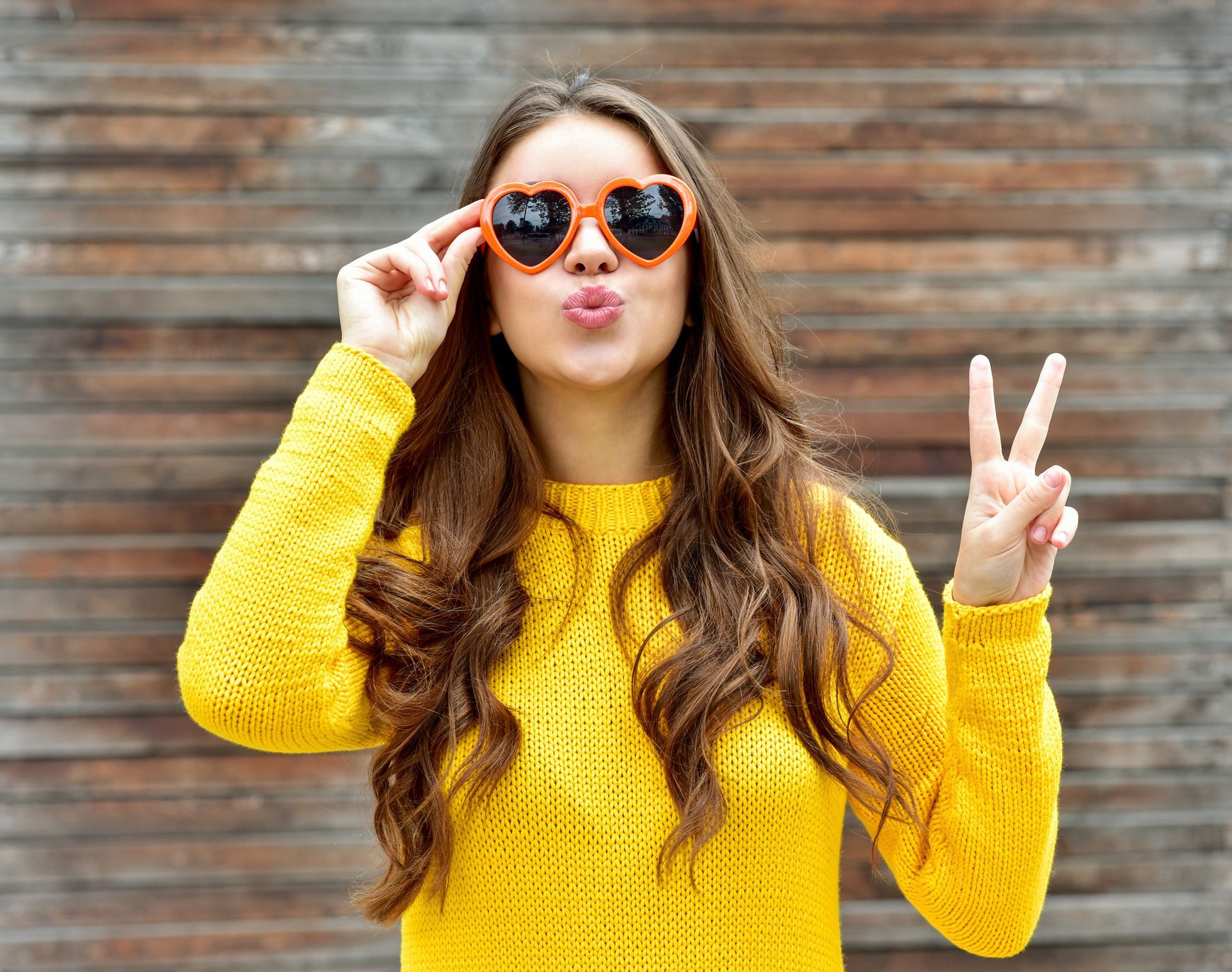 Woman in yellow sweater and heart-shaped sunglasses making a peace sign and blowing a kiss.