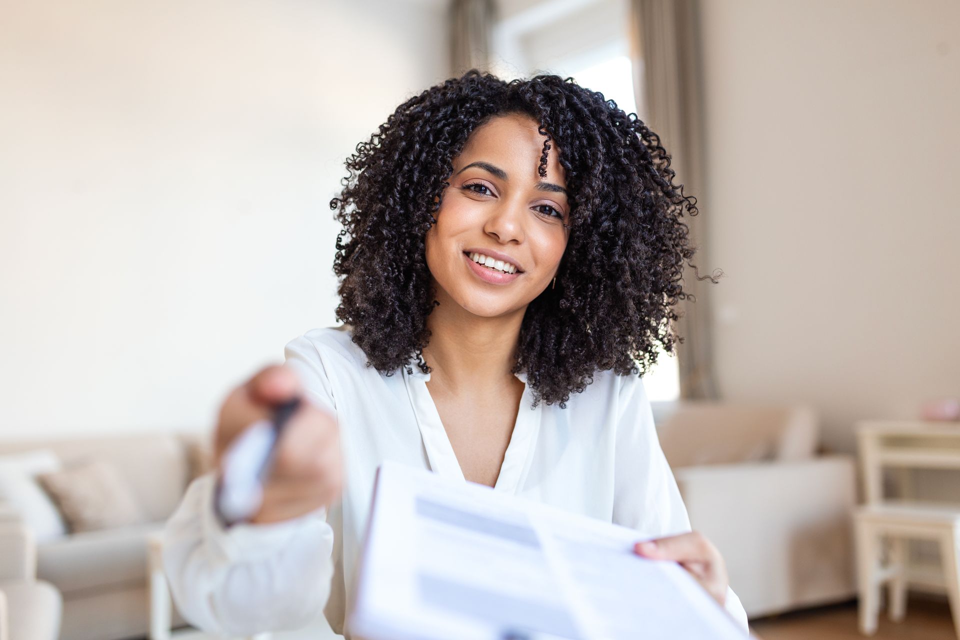 A person with dark, curly hair wearing a white shirt holds out a document and a pen toward the viewer.