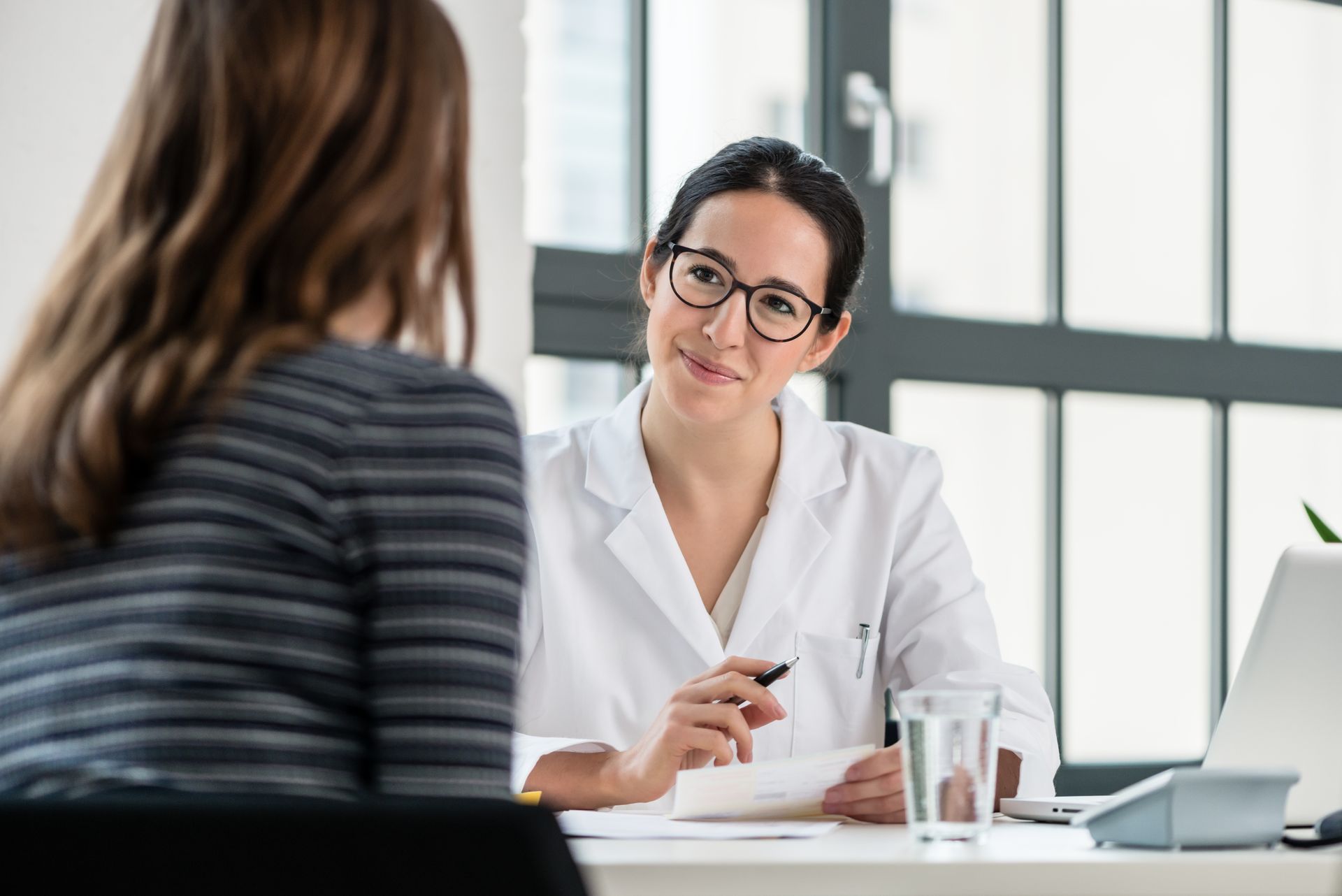 A professional with a white coat consults with a patient across a desk in a bright office.