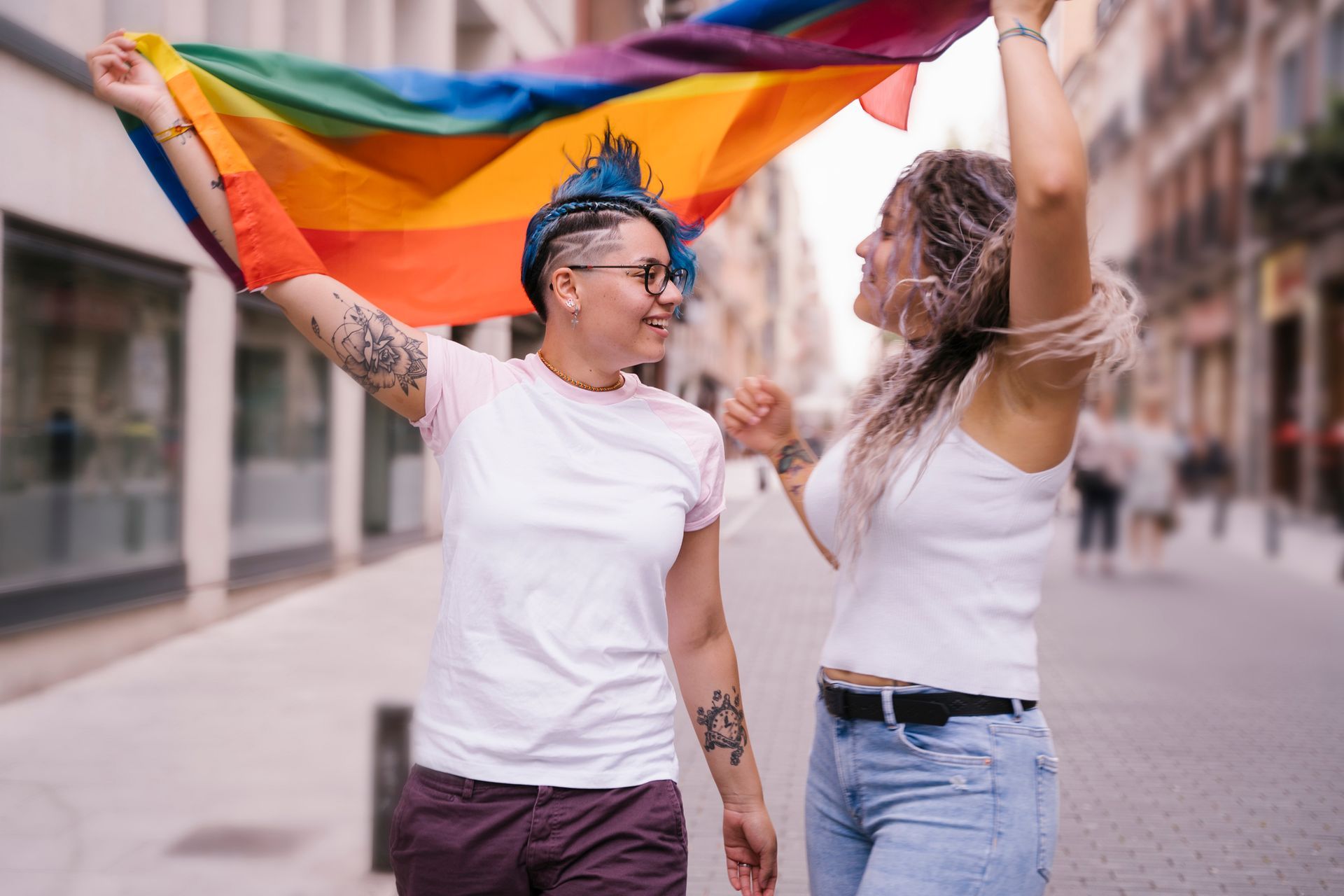 Two people holding a rainbow flag, smiling, walking in a city.