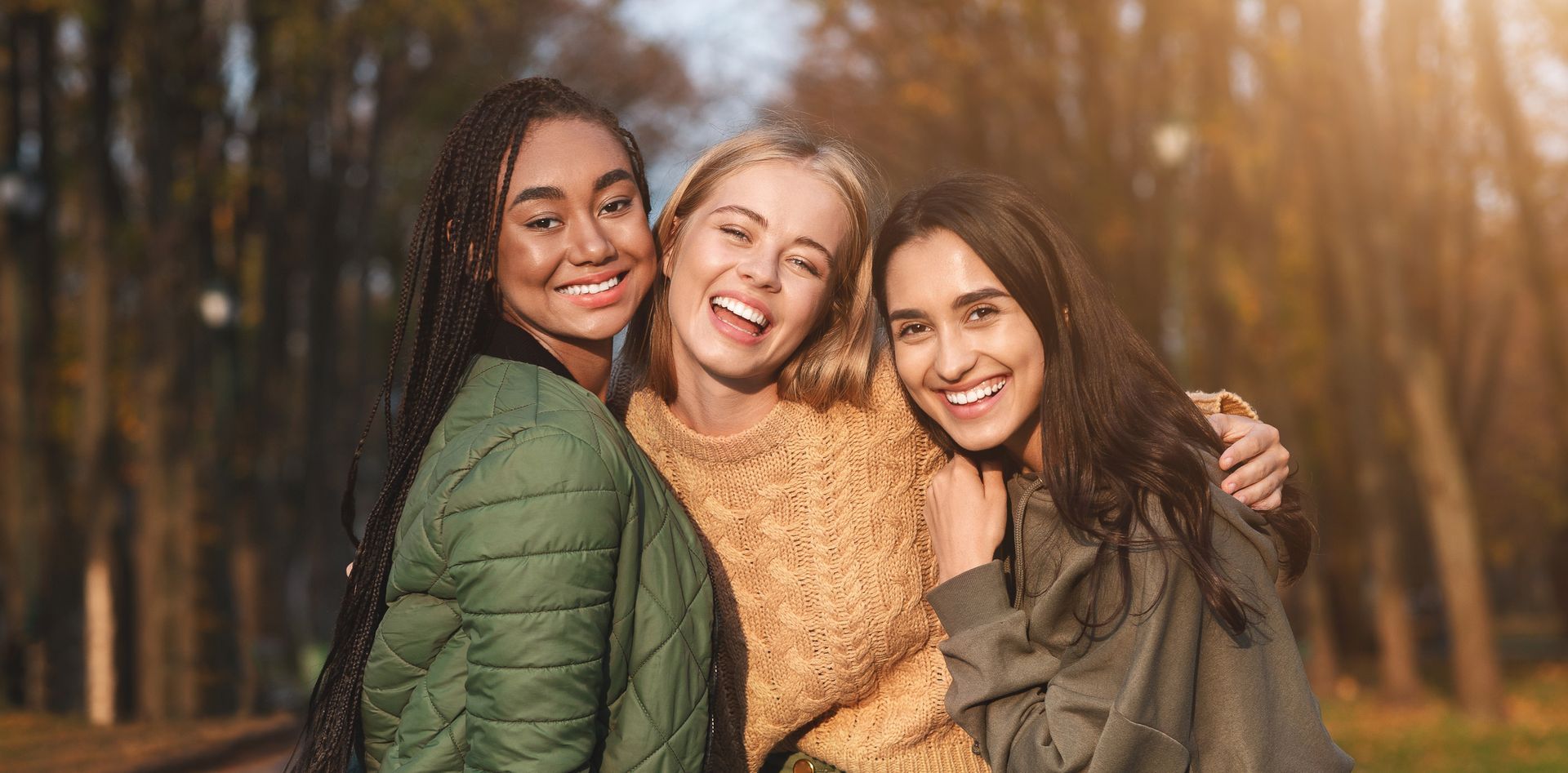 Three smiling friends embrace outdoors with fall foliage.