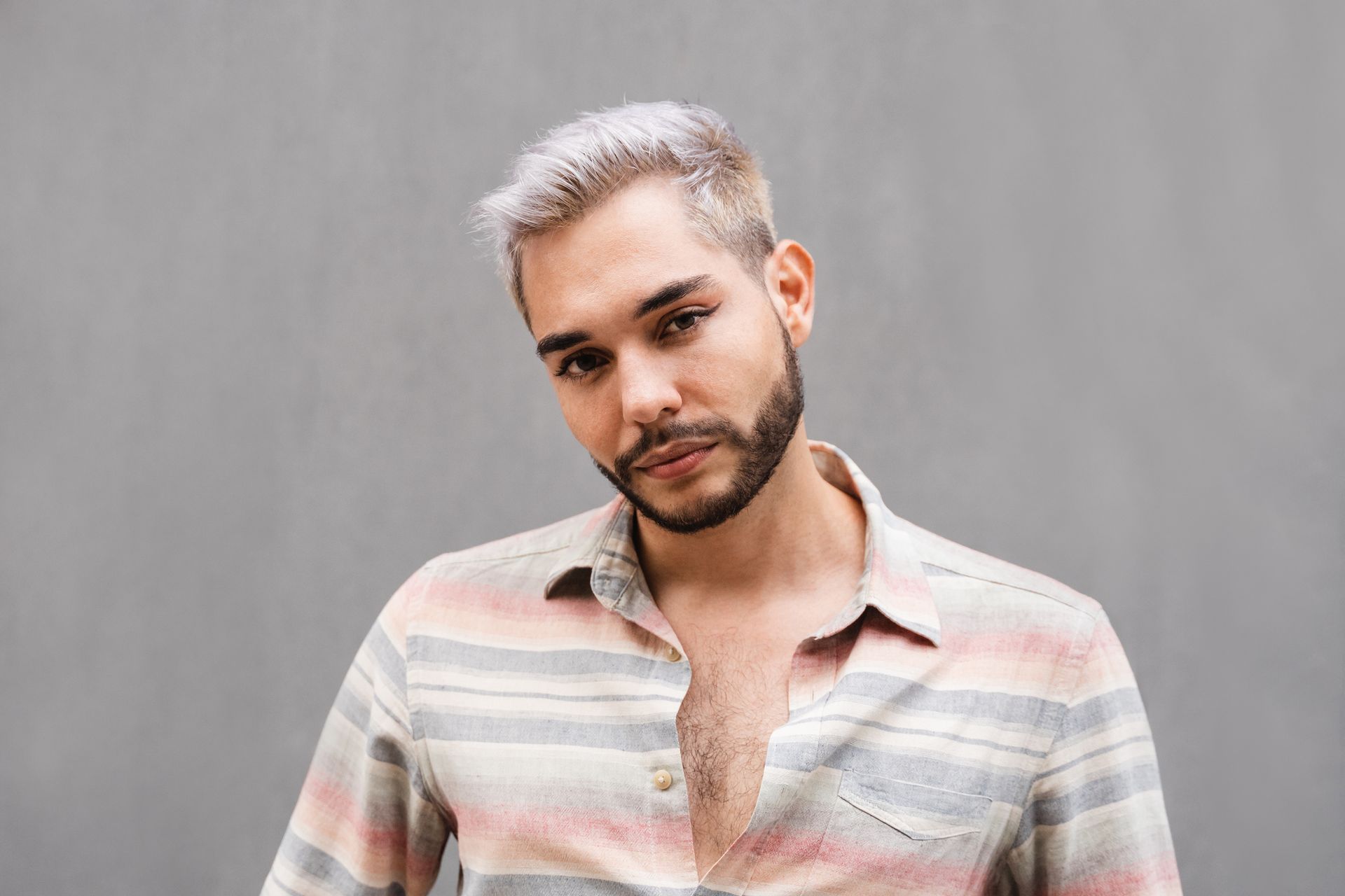 Man with short, light-colored hair and beard, wearing a striped shirt, looking at the camera. Against a gray wall.