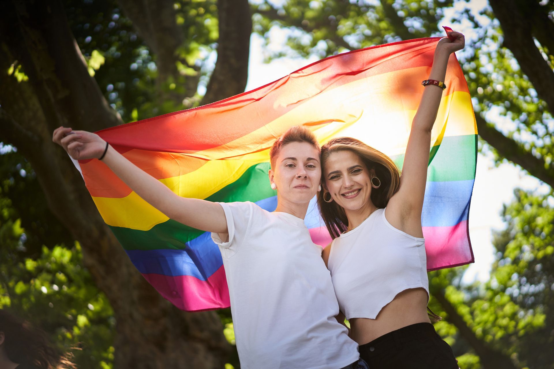 Two people holding up a rainbow flag outdoors, smiling.