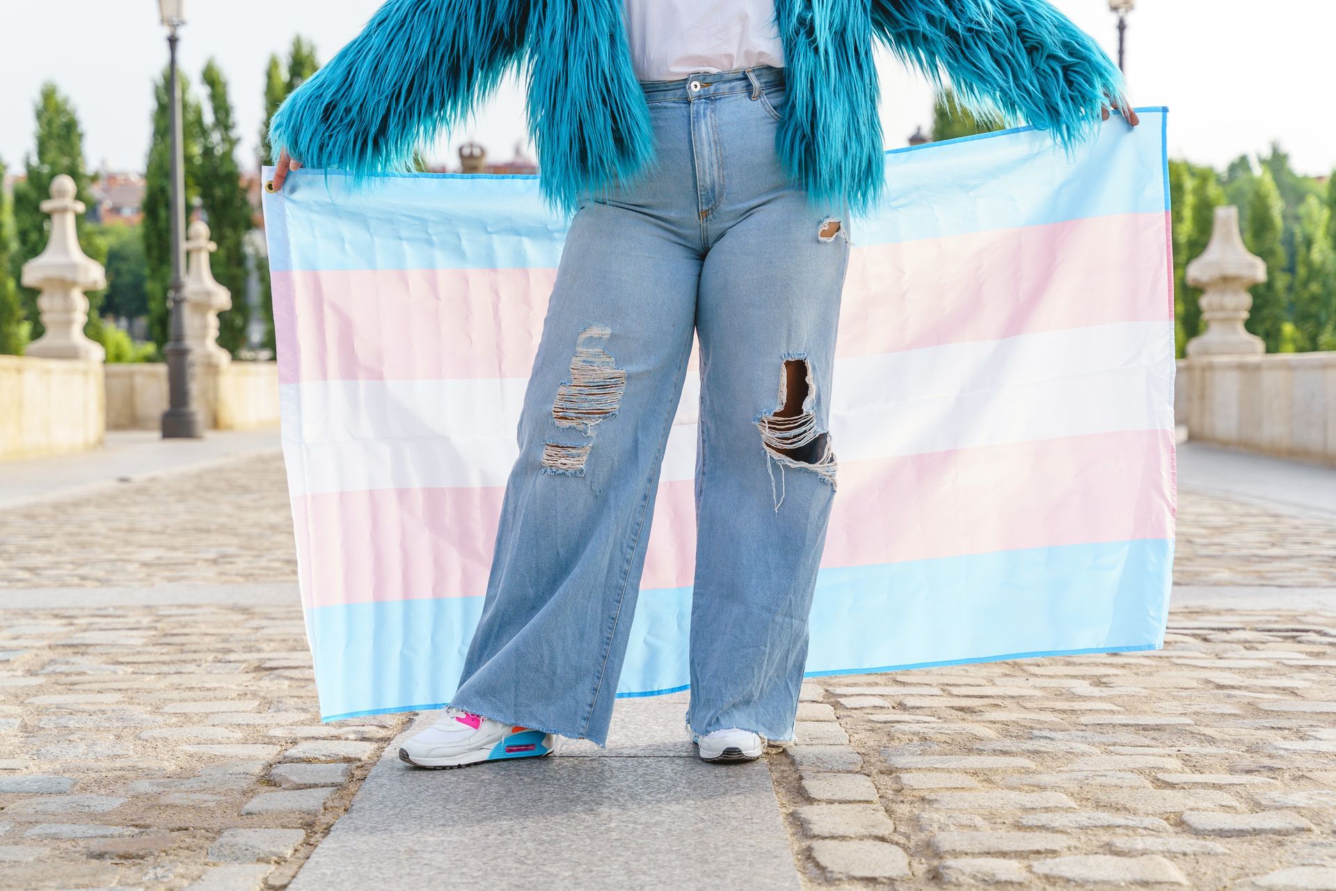 Person holding the transgender pride flag, wearing a blue jacket and ripped jeans. Outdoors.